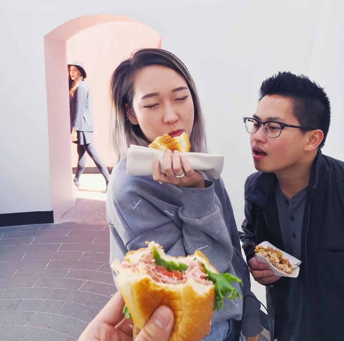 Three people stand together; one woman enjoys a sandwich, a man looks at her food, and another woman walks in the background. A hand holding a sandwich is visible in the foreground.