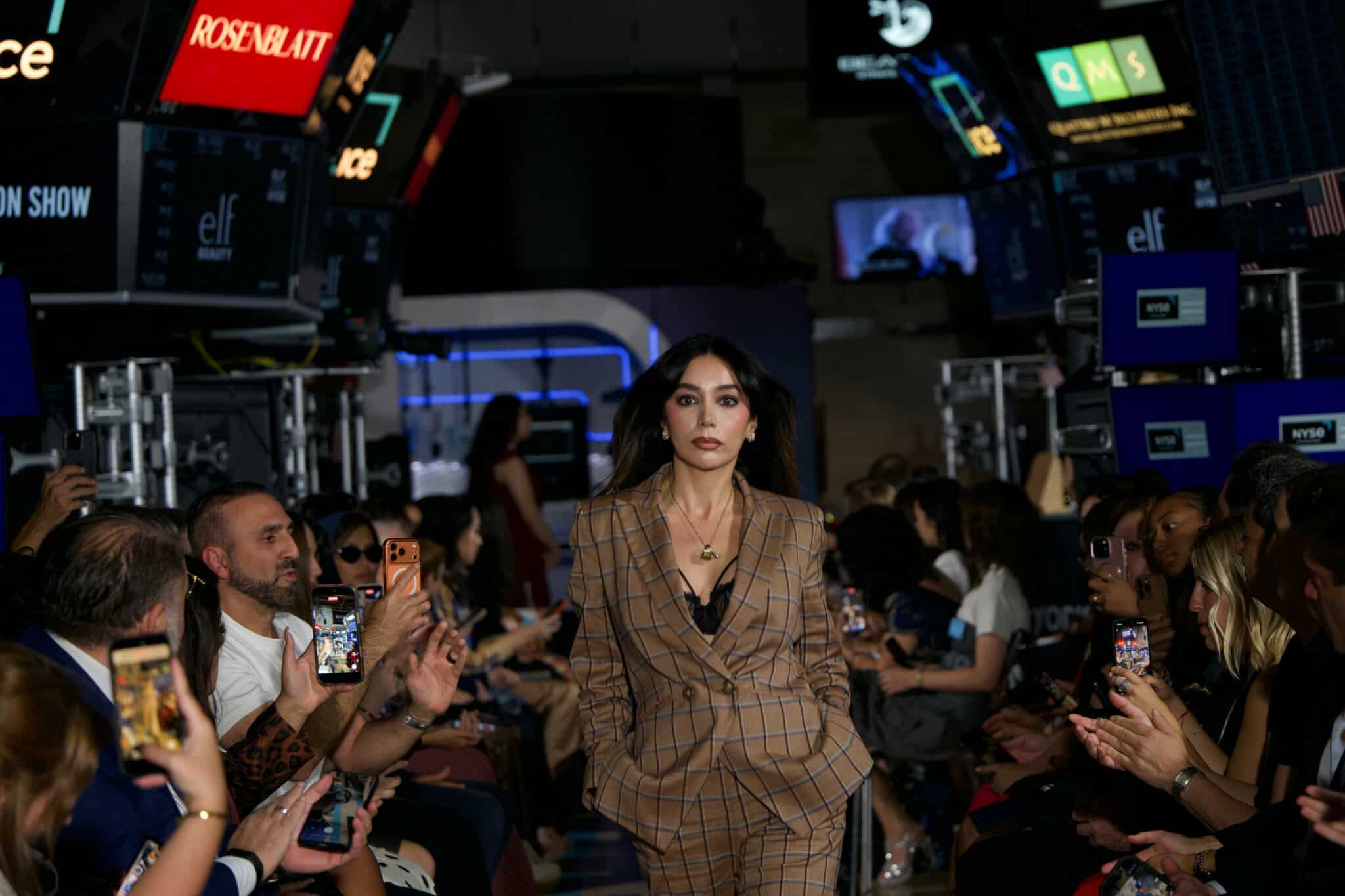 A model in a brown plaid suit walks a runway surrounded by seated spectators, many holding up phones to take photos; digital screens and lights are visible above the audience.