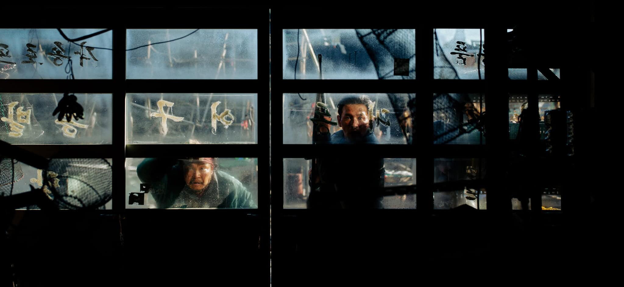 Two people peer intensely through a foggy, windowed garage door. Light and shadows highlight their faces, with rusty objects and markings visible on the glass. The scene feels tense and mysterious.