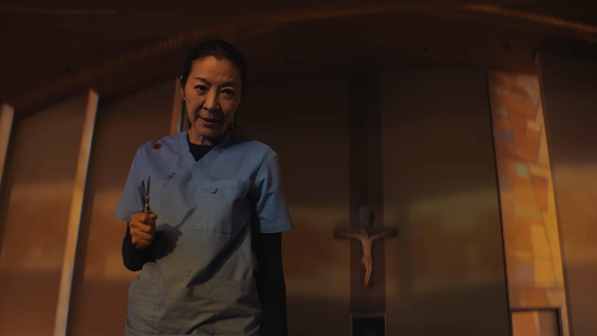A woman in blue scrubs holding scissors stands in a dimly lit church, with a crucifix and stained glass in the background. She has blood stains on her clothes and a serious expression on her face.