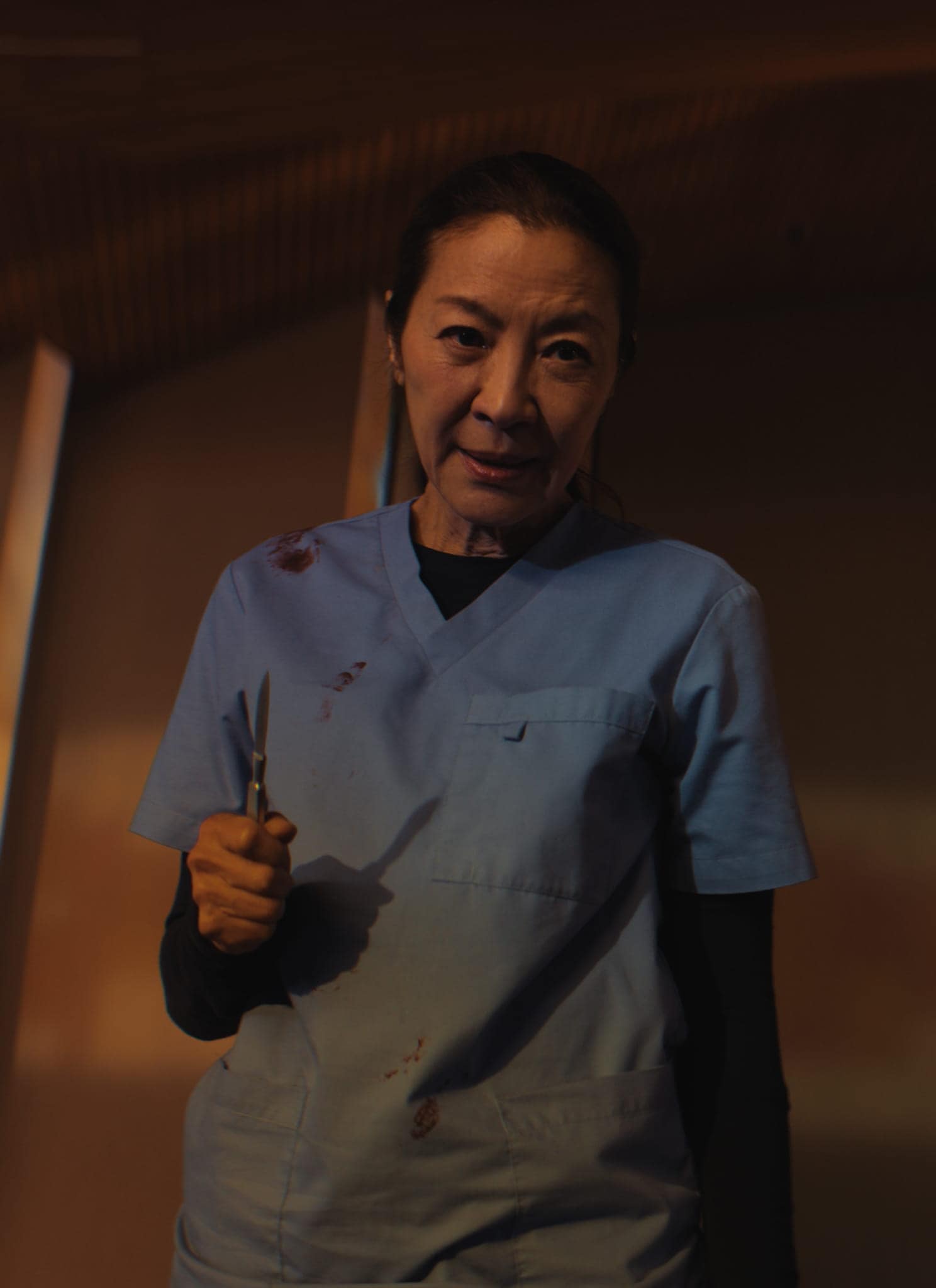 A woman in blue scrubs holding scissors stands in a dimly lit church, with a crucifix and stained glass in the background. She has blood stains on her clothes and a serious expression on her face.