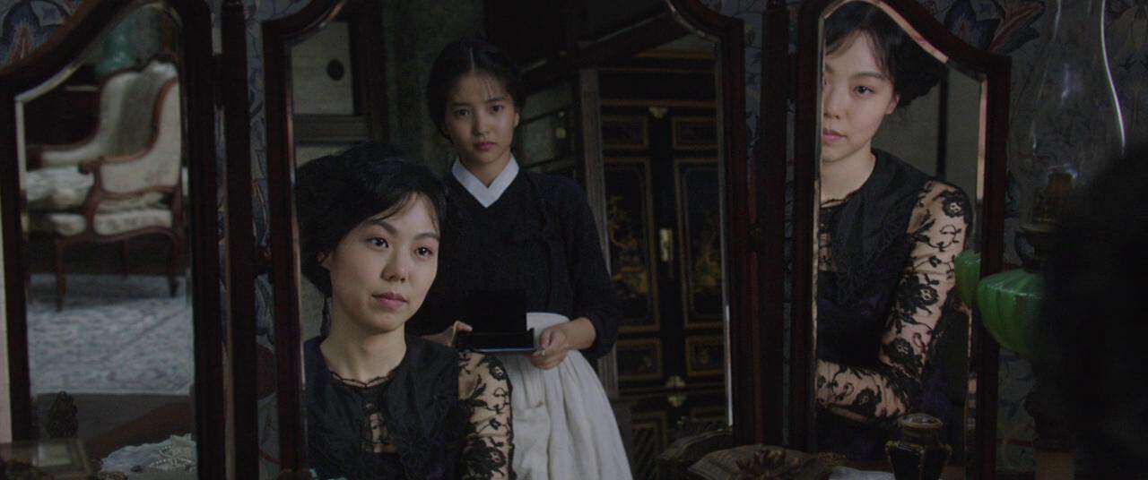 A woman with dark hair in a black lace-sleeved dress sits before a vanity mirror while another woman in a black top and white apron stands behind her, their reflections visible in the mirror panels.