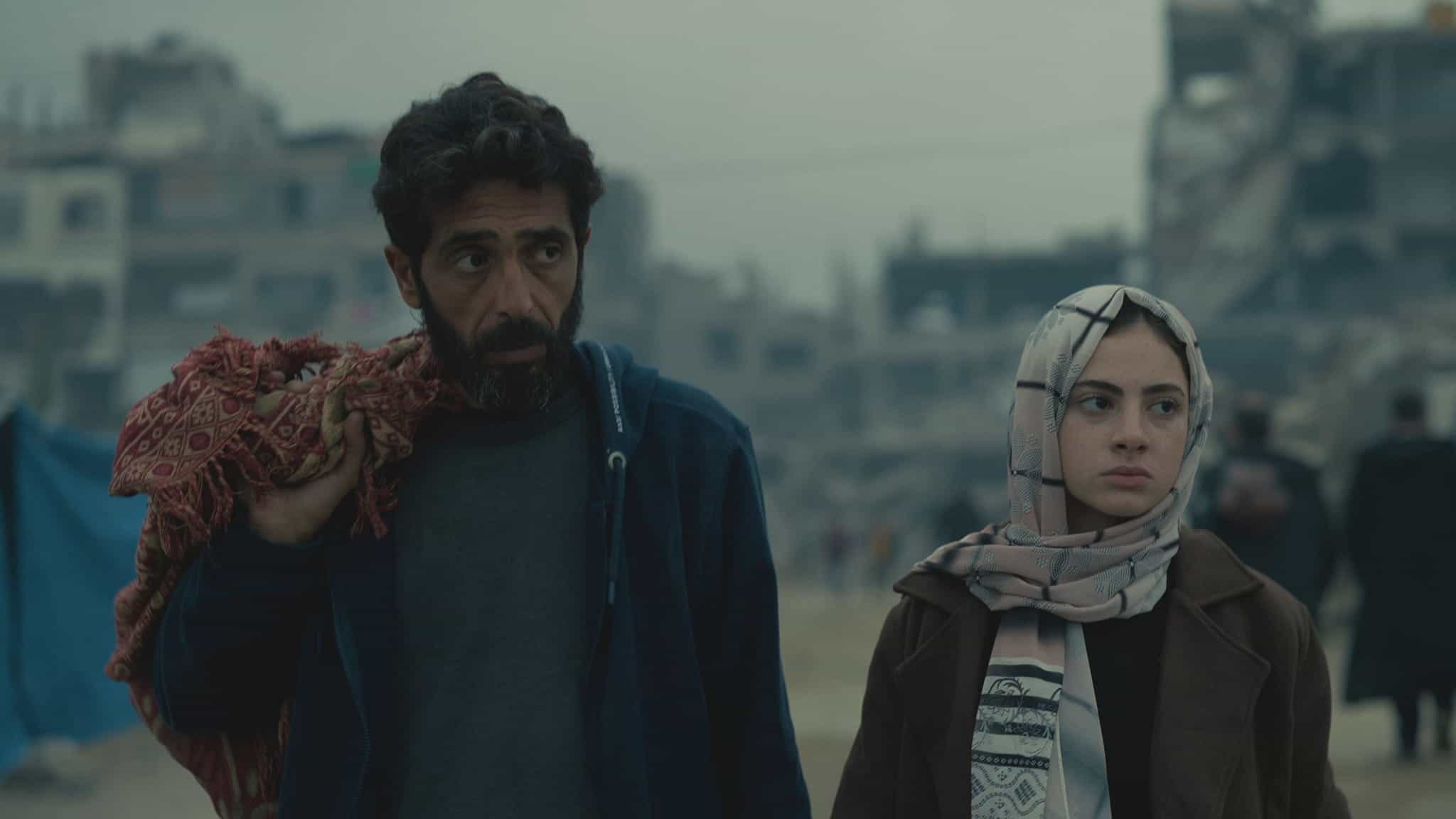 A man and a young woman stand side by side in front of a war-torn, grey urban landscape. The man carries a bundle over his shoulder; the woman wears a patterned headscarf and coat. Both look serious and concerned.