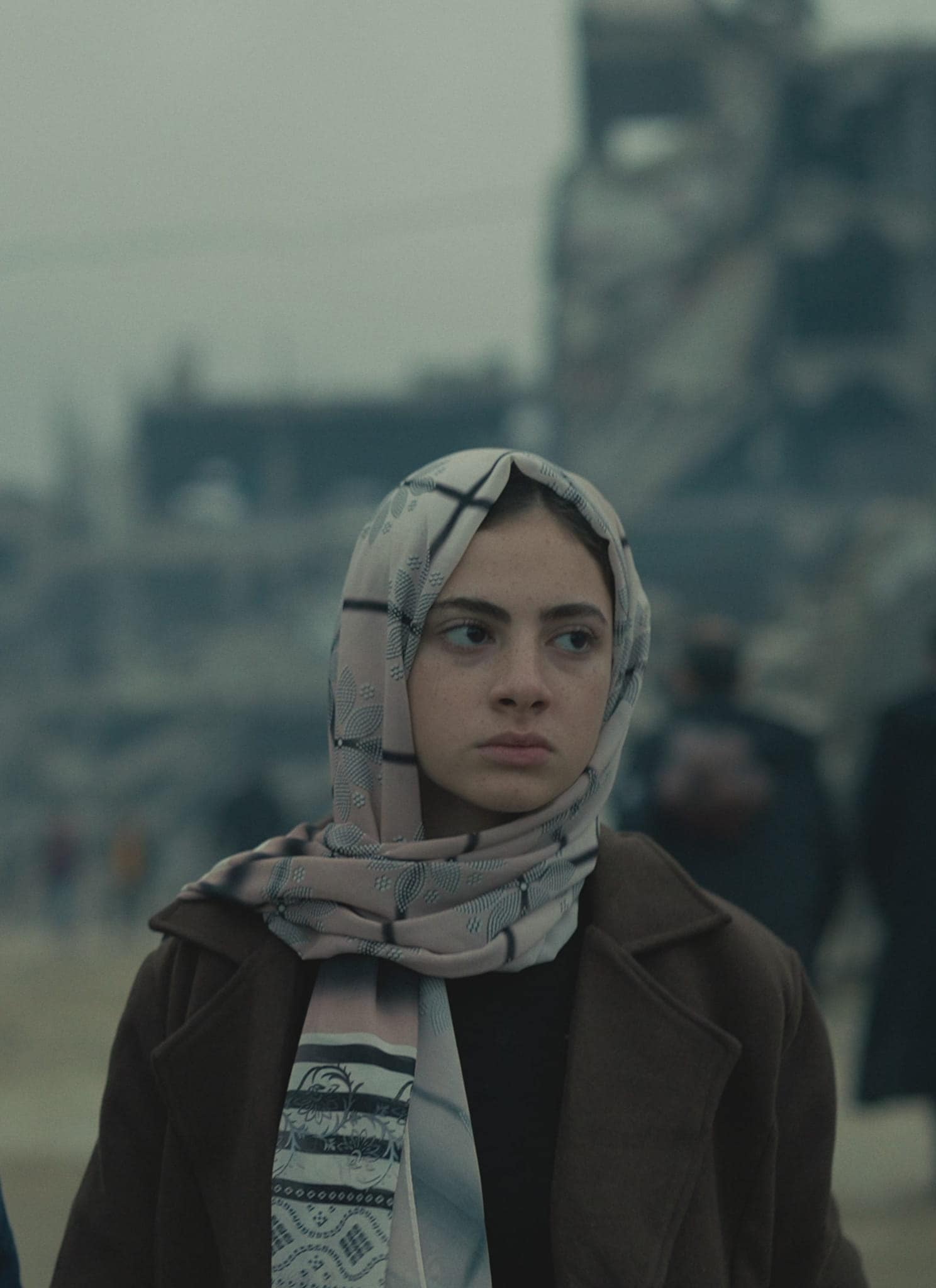 A man and a young woman stand side by side in front of a war-torn, grey urban landscape. The man carries a bundle over his shoulder; the woman wears a patterned headscarf and coat. Both look serious and concerned.