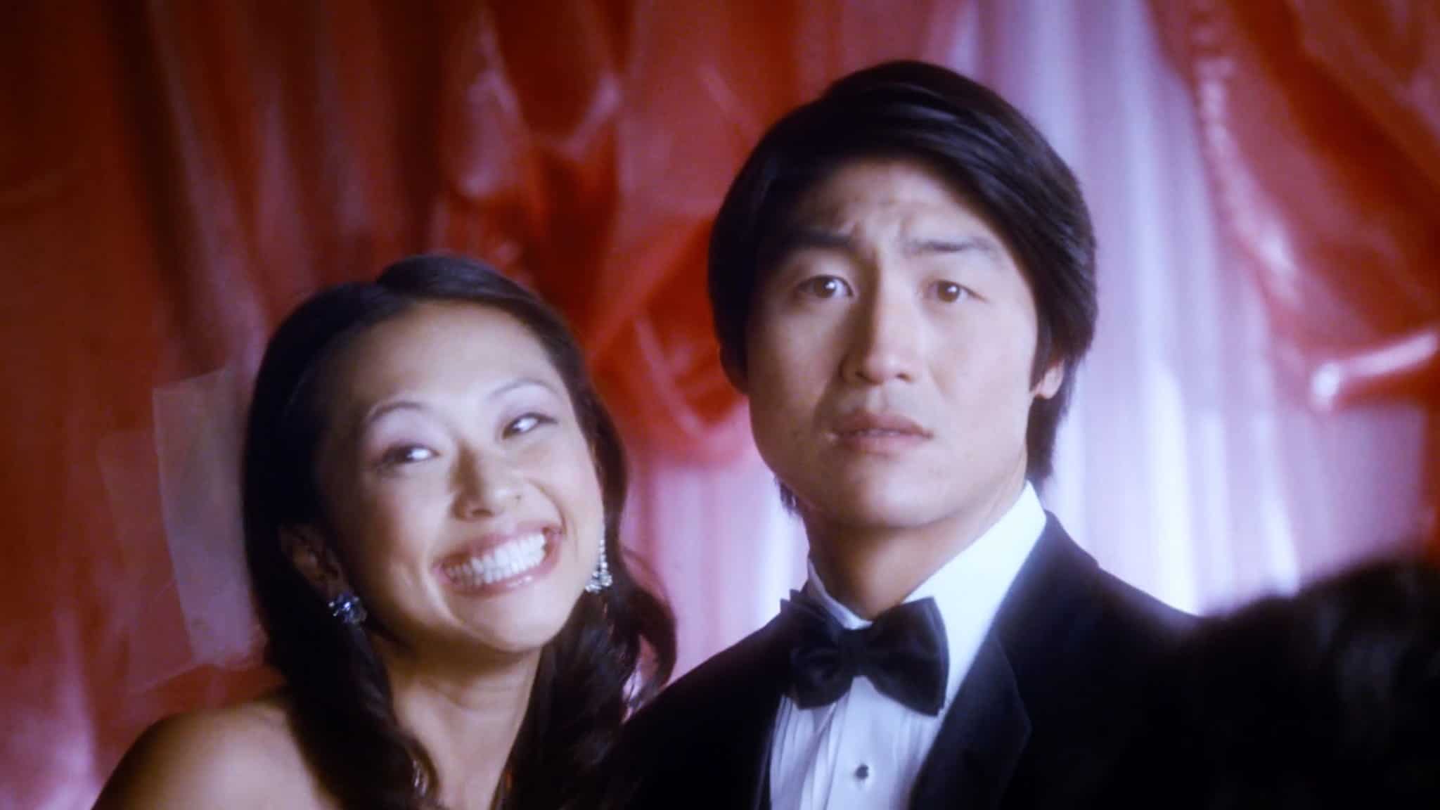 A woman in a dress smiles widely, while a man beside her in a tuxedo looks serious. Behind them, a red and pink backdrop evokes the elegance of a wedding palace.