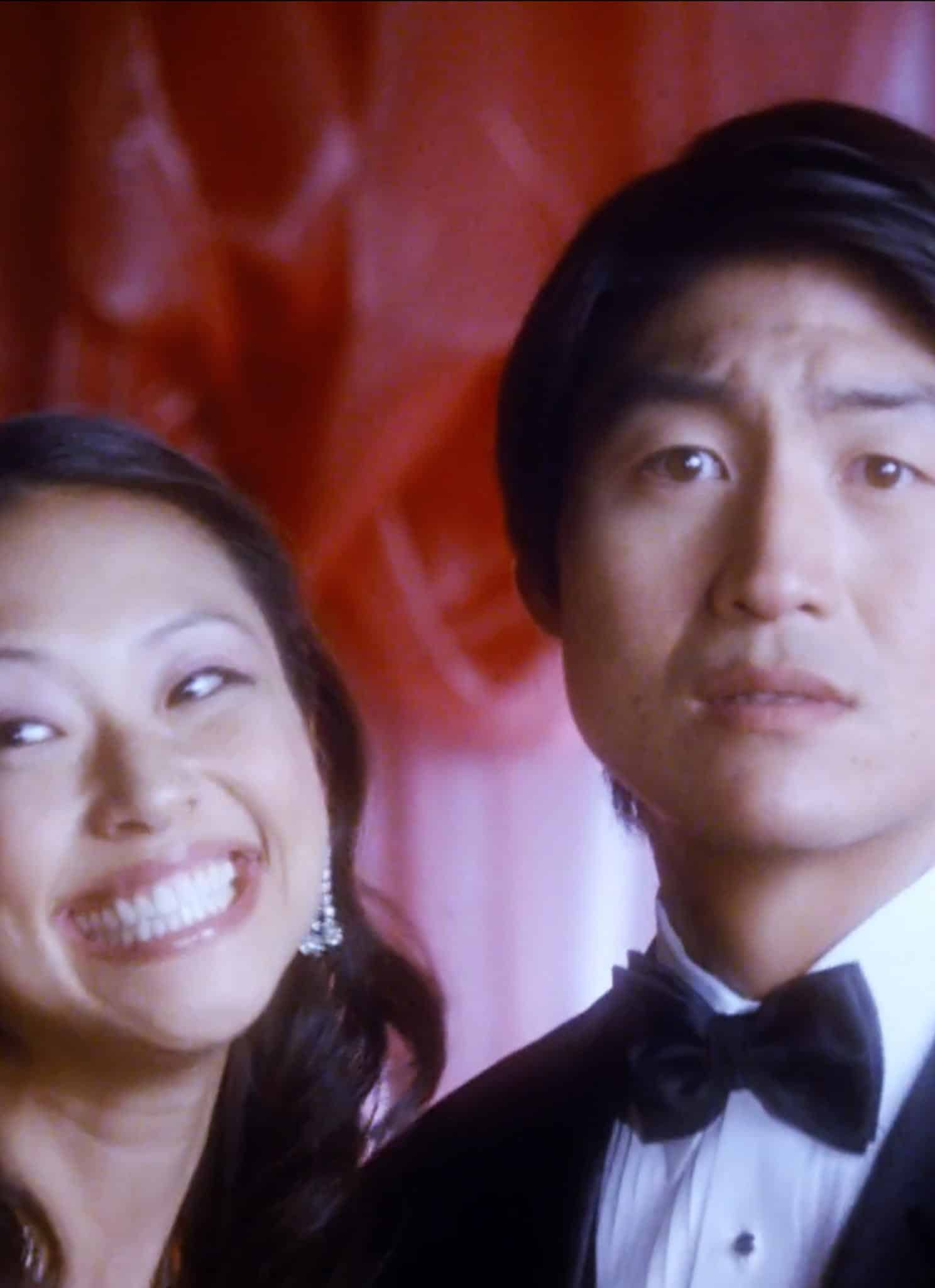 A woman in a dress smiles widely, while a man beside her in a tuxedo looks serious. Behind them, a red and pink backdrop evokes the elegance of a wedding palace.