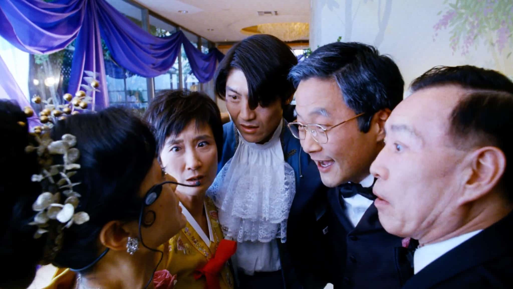 Five people in formal attire cluster closely together indoors, looking intently at the woman on the left, who is holding a monocle. Purple drapes and large windows—reminiscent of a grand wedding palace—are visible in the background.
