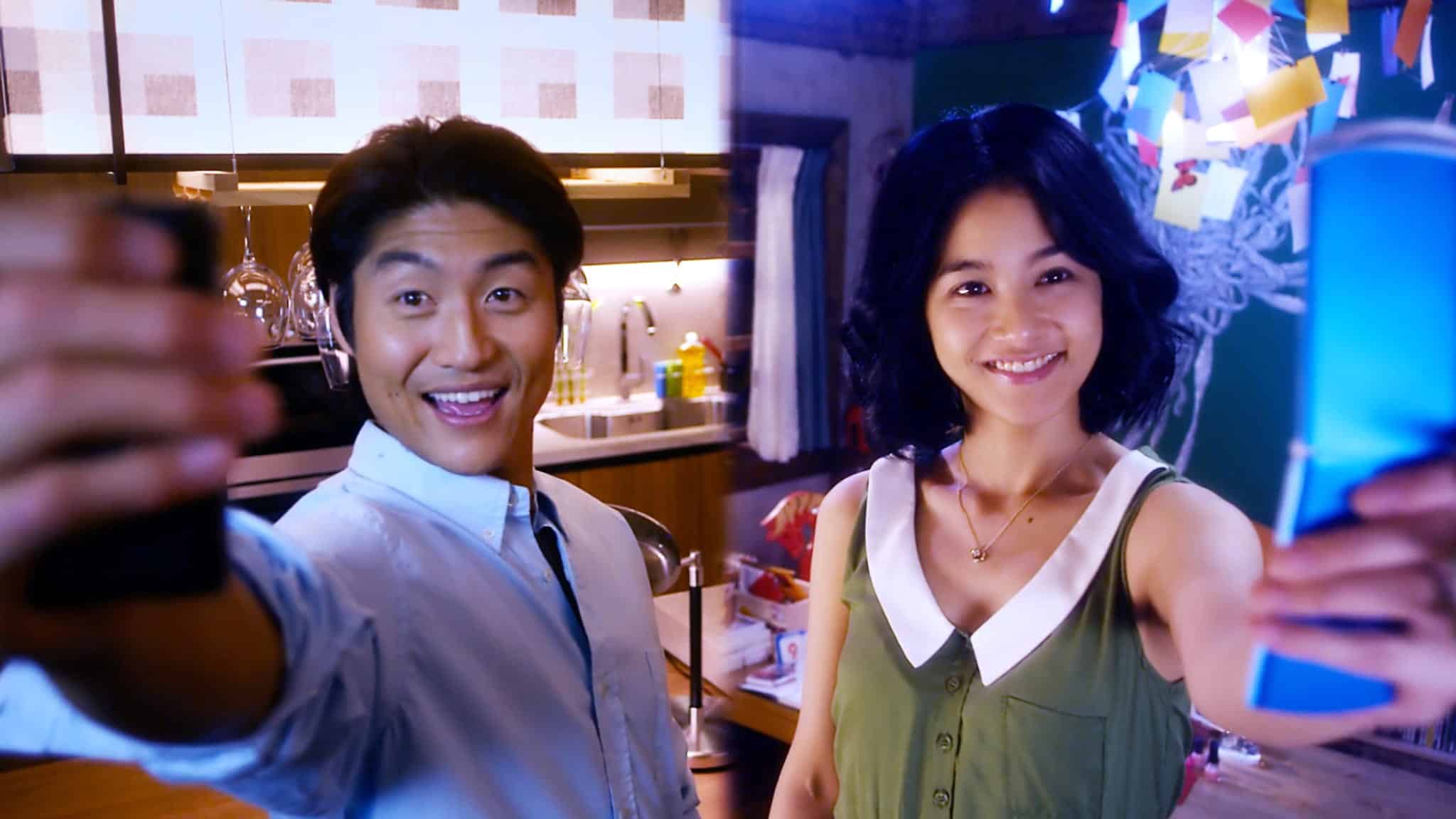 A man and woman smile and hold up drinks while taking a selfie in a brightly lit kitchen, decorated with lights and colorful paper shapes that transform the space into their own cozy wedding palace.