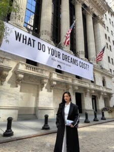 A woman in a black and white coat stands outside a large building with American flags and a banner reading WHAT DO YOUR DREAMS COST? RISE FASHION SHOW..