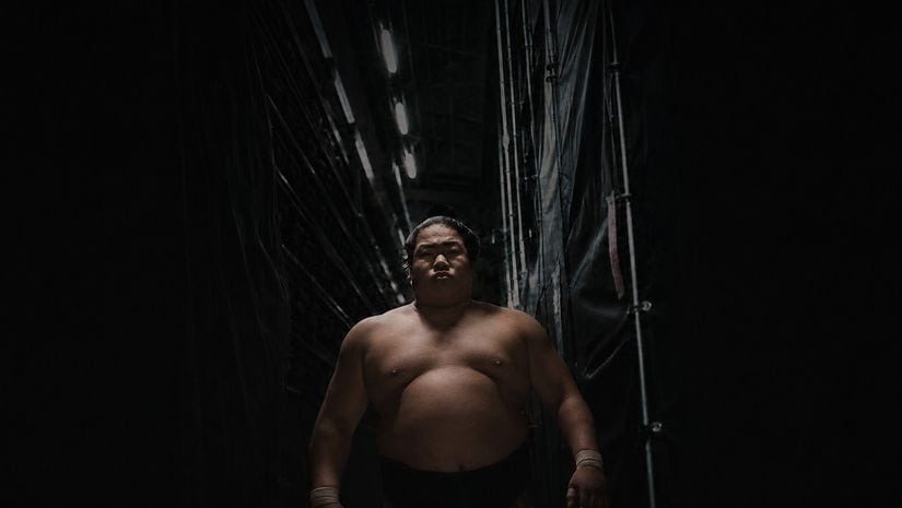 A sumo wrestler stands shirtless in a dark corridor with lights above, looking forward with a serious expression.