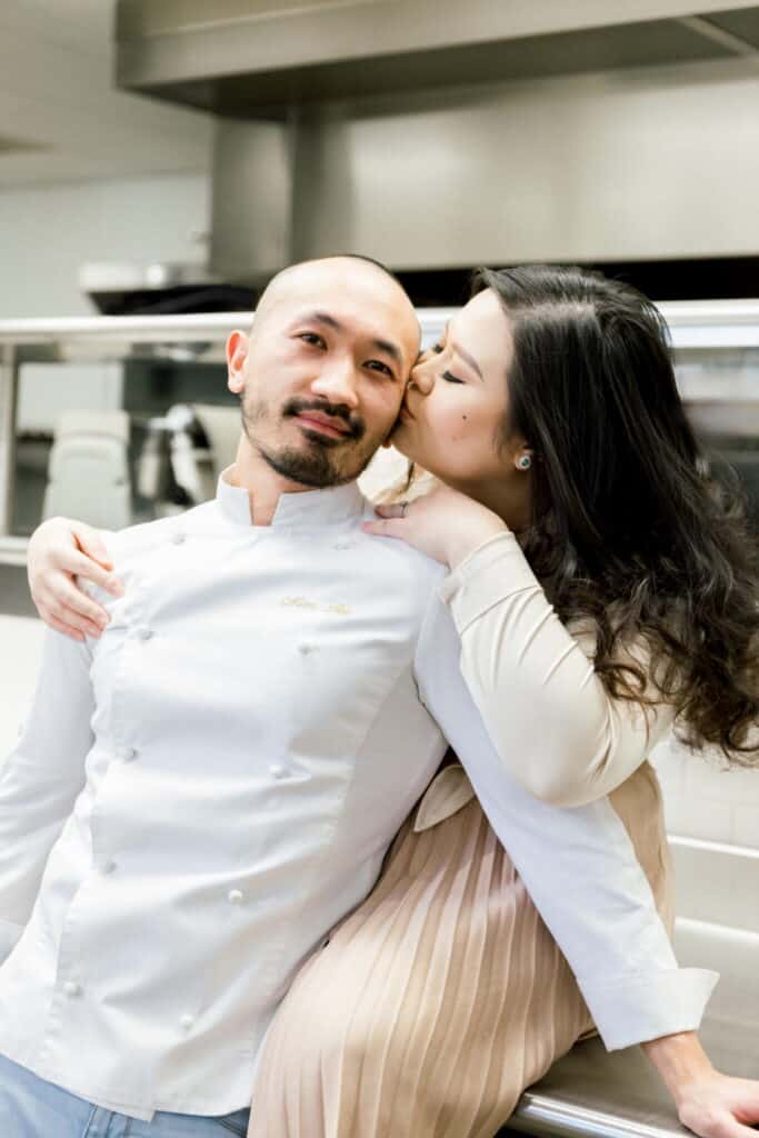 A woman kisses a smiling man on the temple in a modern kitchen. The man wears a white chefs coat, while the woman is dressed in light colors and sits close with her arm around his shoulders.