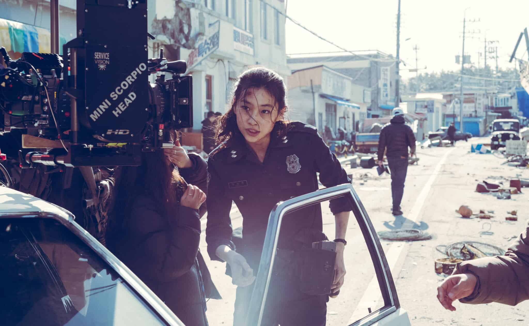 A woman in a police uniform steps out of a car on a debris-strewn street, with a film crew and camera nearby, during the shooting of a dramatic scene reminiscent of Na Hong-jin’s intense cinematic style.