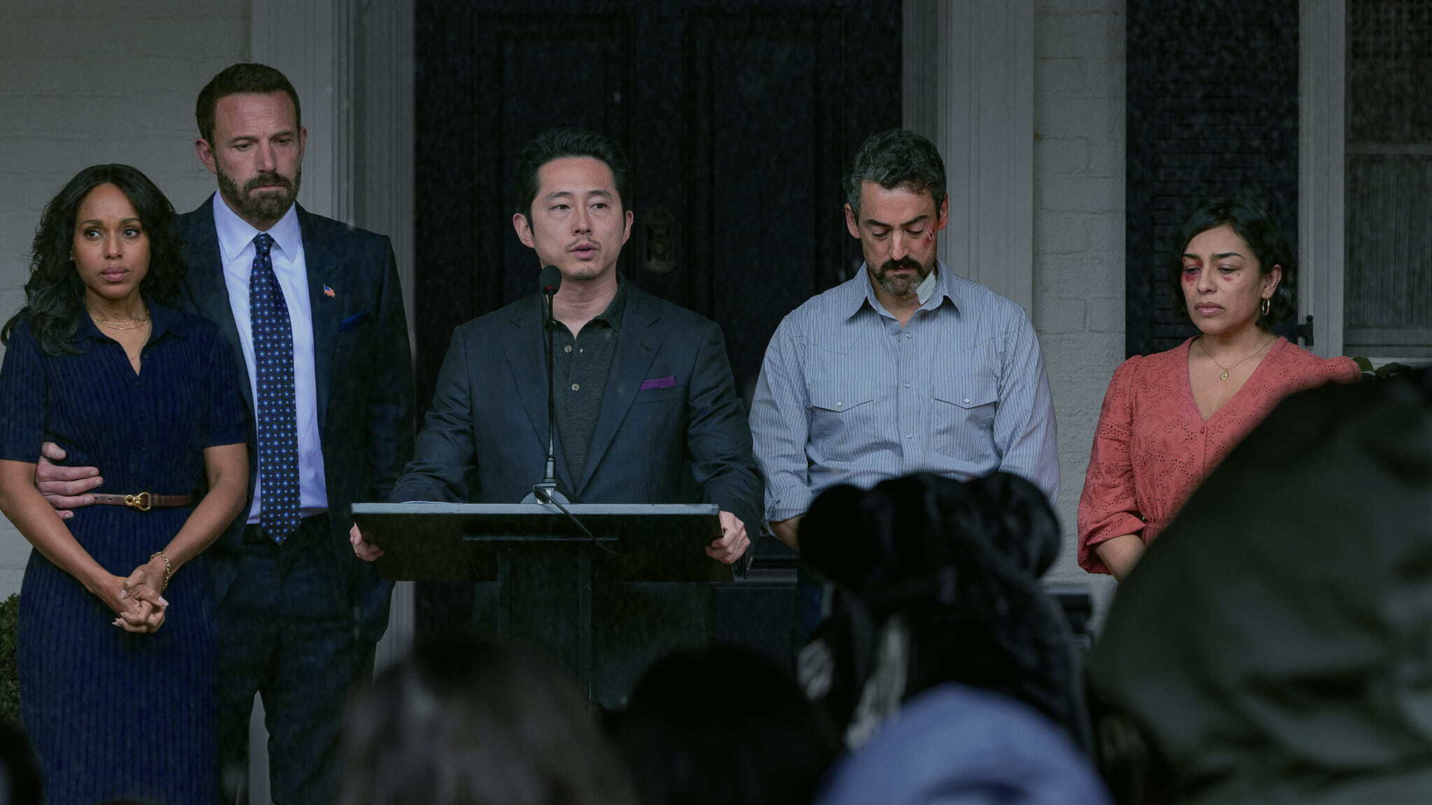 Five people stand in a line during a press conference; the man in the center speaks at a podium while the others stand behind him, some appearing injured.