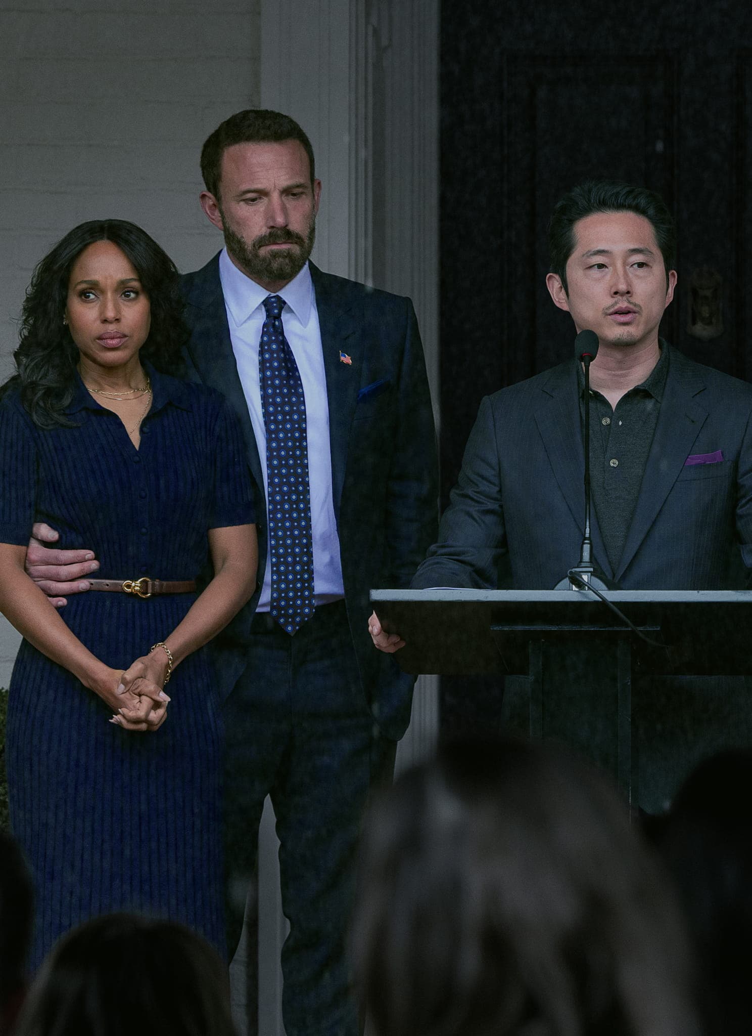 Five people stand in a line during a press conference; the man in the center speaks at a podium while the others stand behind him, some appearing injured.