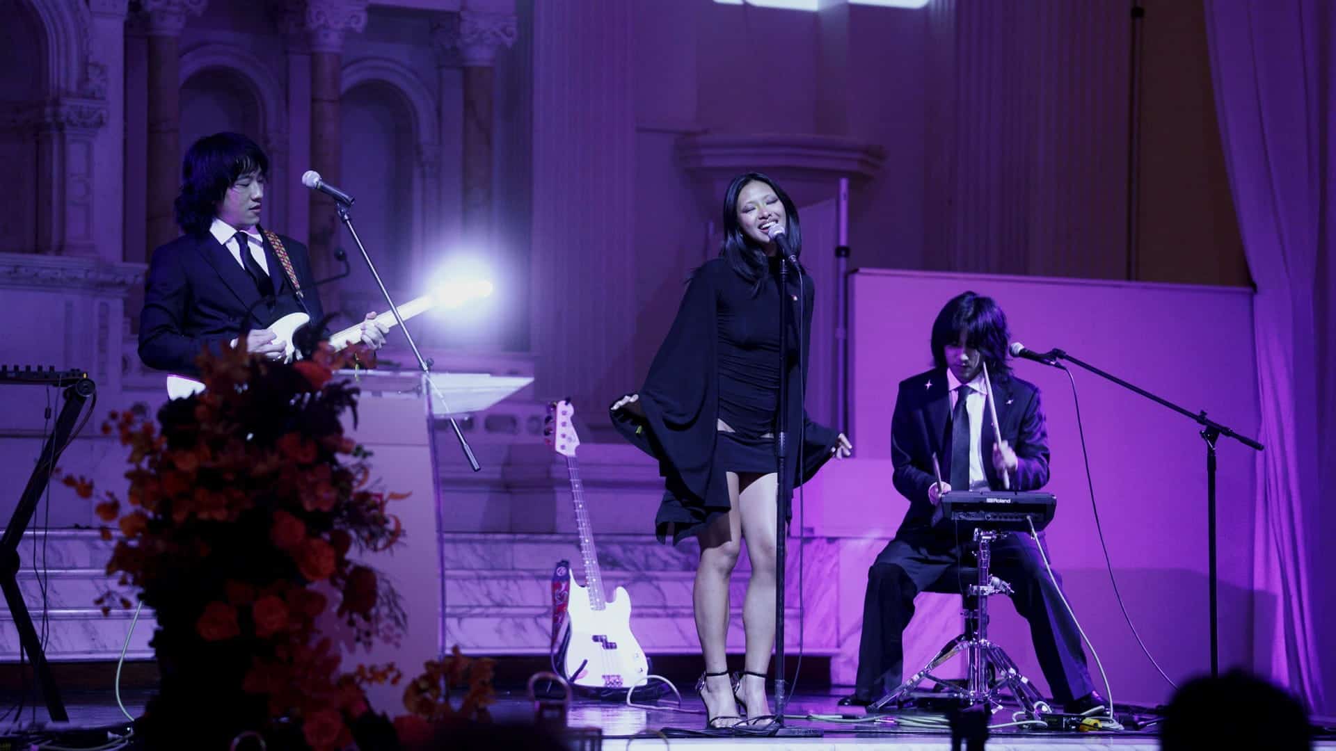 A band performing on stage, with a female singer in a black dress in the center, a guitarist to her left, and a percussionist playing a digital drum pad to her right.