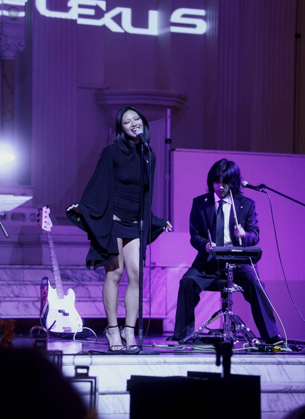A band performing on stage, with a female singer in a black dress in the center, a guitarist to her left, and a percussionist playing a digital drum pad to her right.