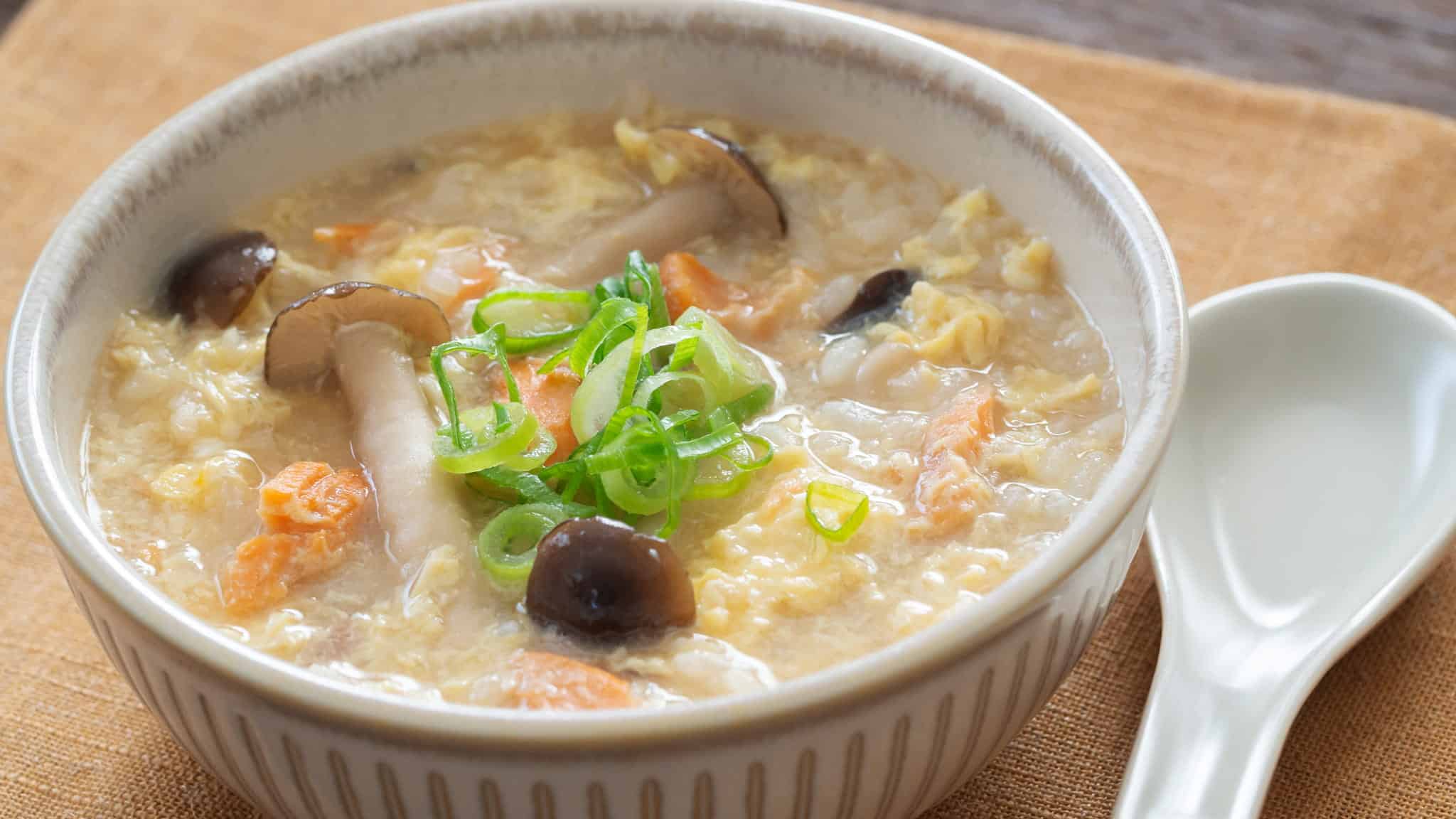 A bowl of egg drop soup with mushrooms, carrots, and sliced green onions, served with a white ceramic spoon.