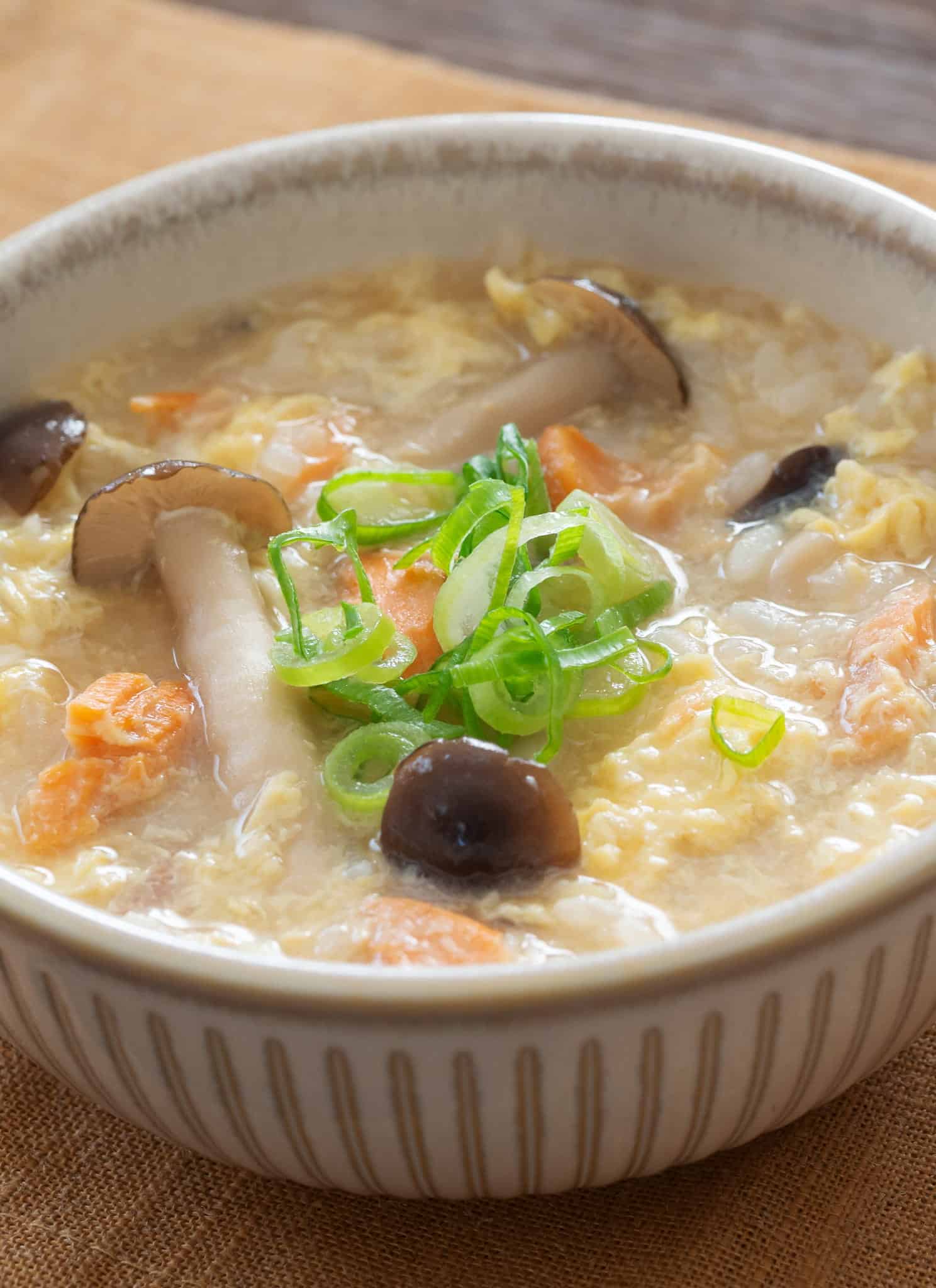 A bowl of egg drop soup with mushrooms, carrots, and sliced green onions, served with a white ceramic spoon.