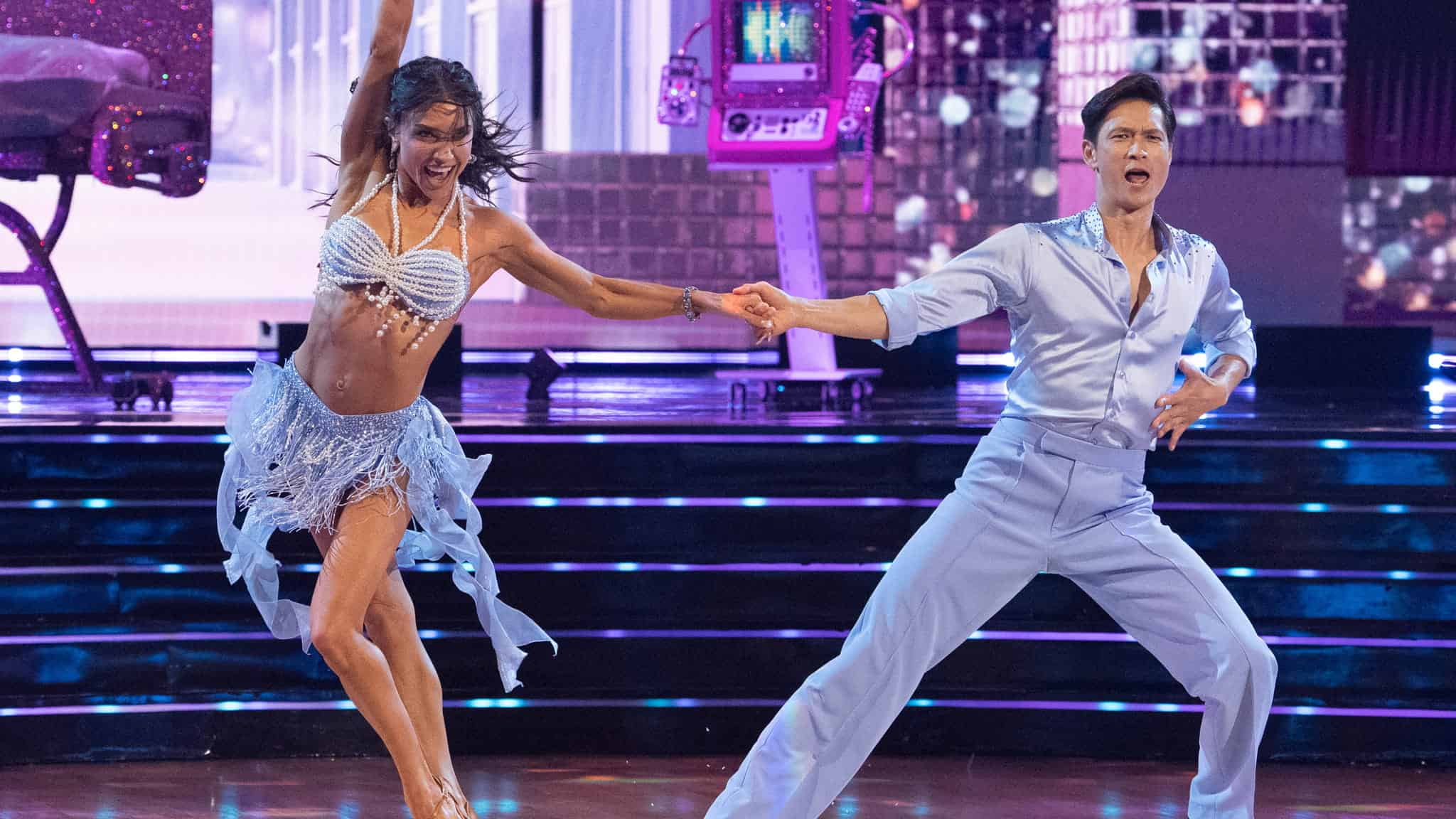 A woman in a beaded blue fringe costume and a man in a light blue button-up shirt perform a dance on a stage.