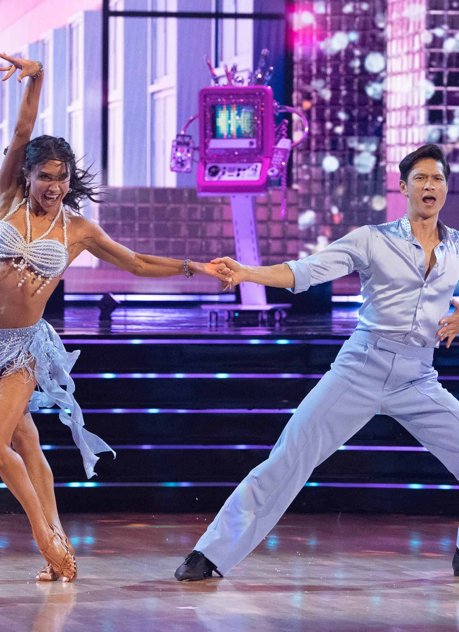 A woman in a beaded blue fringe costume and a man in a light blue button-up shirt perform a dance on a stage.