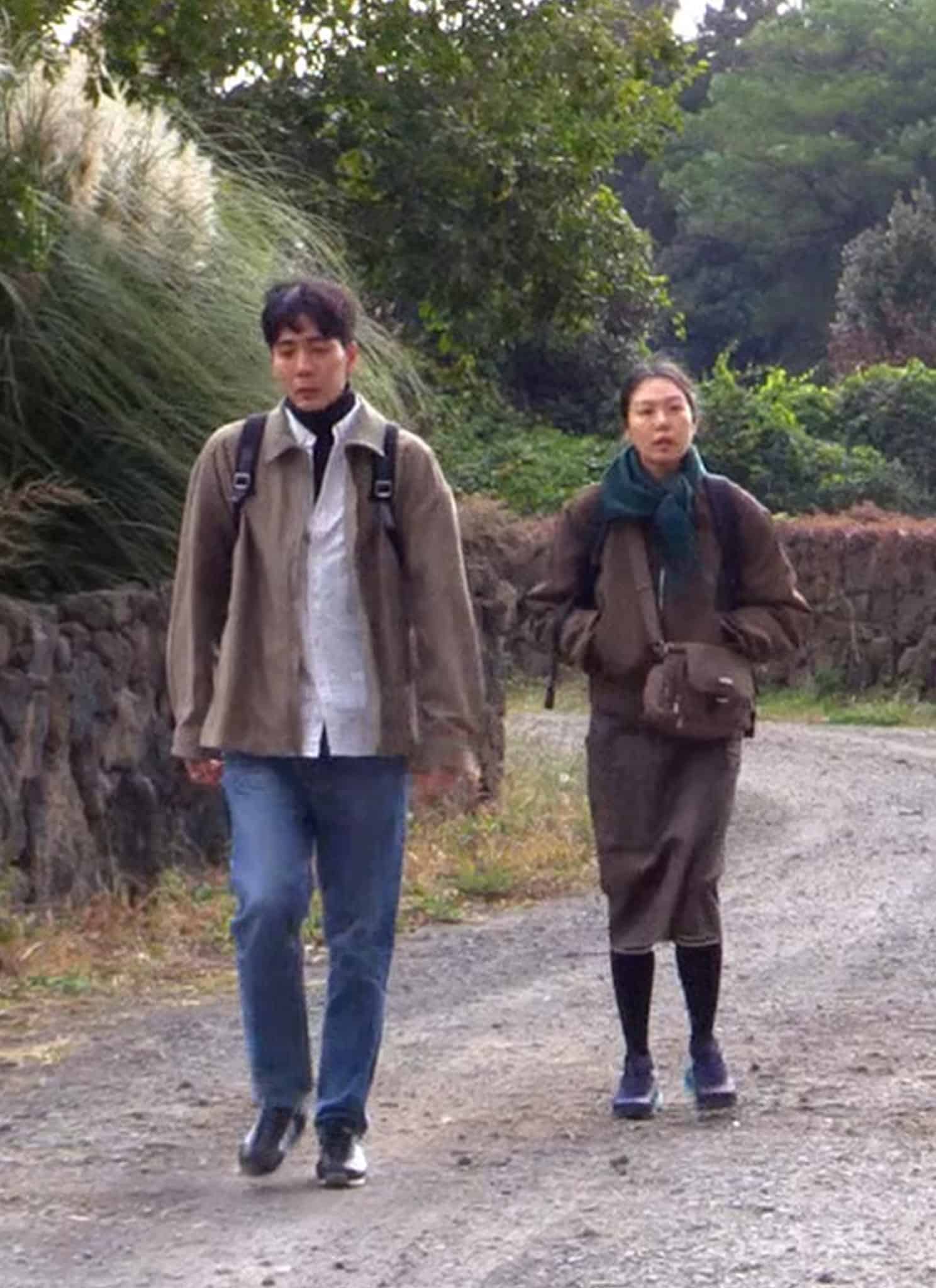 A man and a woman walk side-by-side along a gravel path bordered by a stone wall and tall grasses, in a scene reminiscent of a Hong Sang-soo film.
