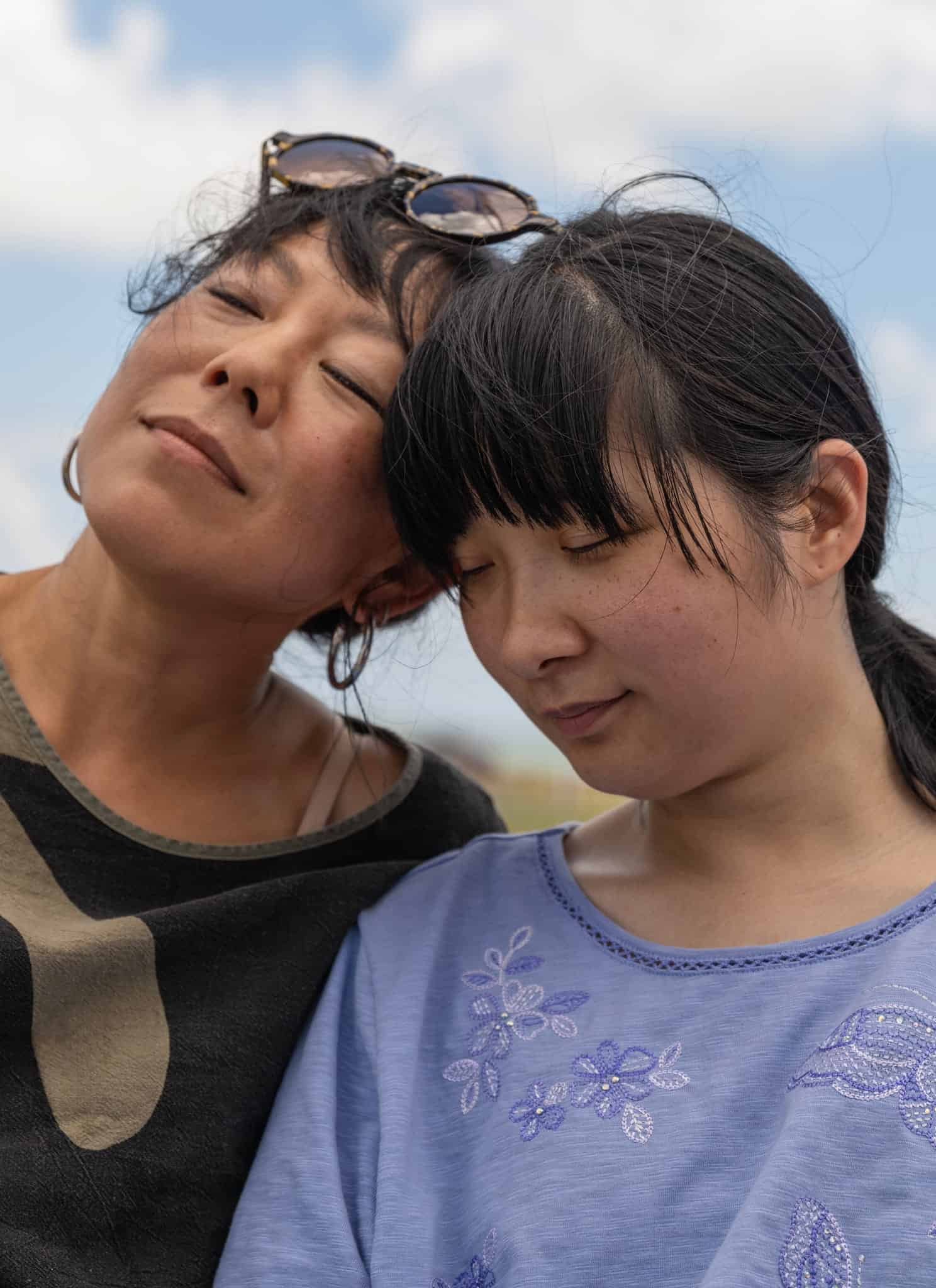 Two women resting their heads against each other outdoors with eyes closed.