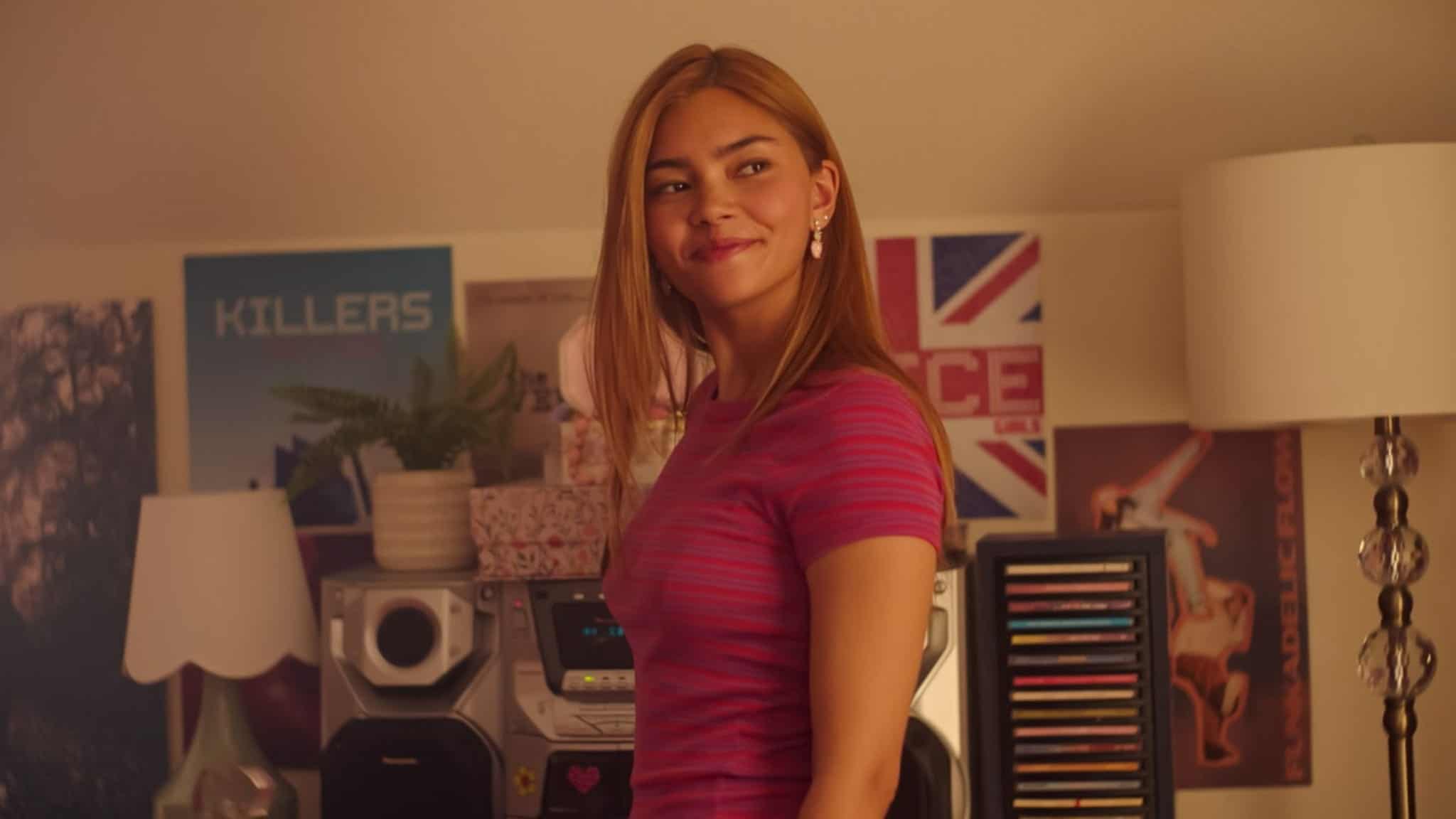 A young woman with long hair wearing a striped pink shirt looks over her shoulder while standing in a room with posters on the wall.