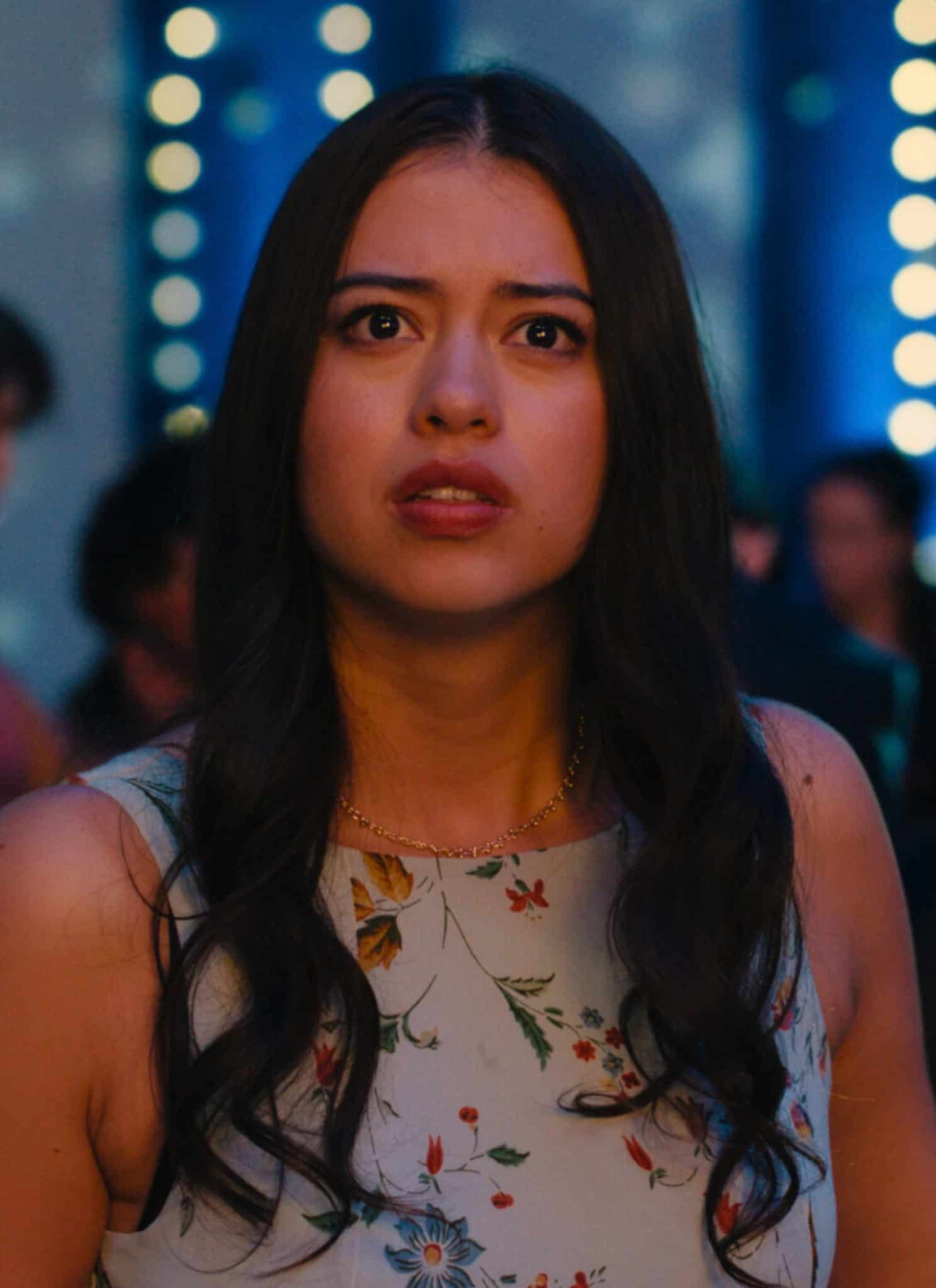 A young woman with a concerned expression stands in a crowd under blue string lights.