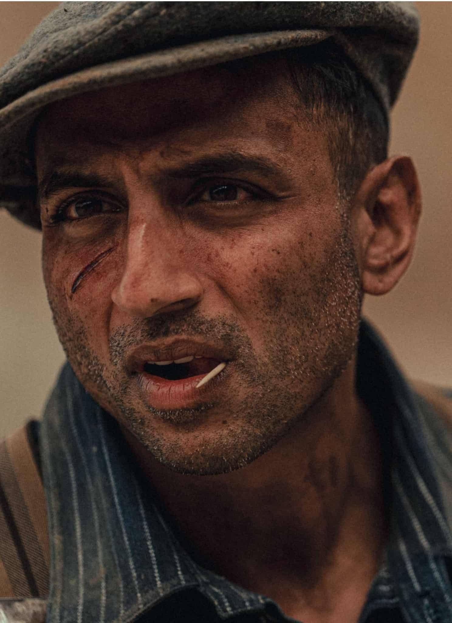 Close-up of a man in a flat cap with a small cut on his cheek and a toothpick in his mouth.