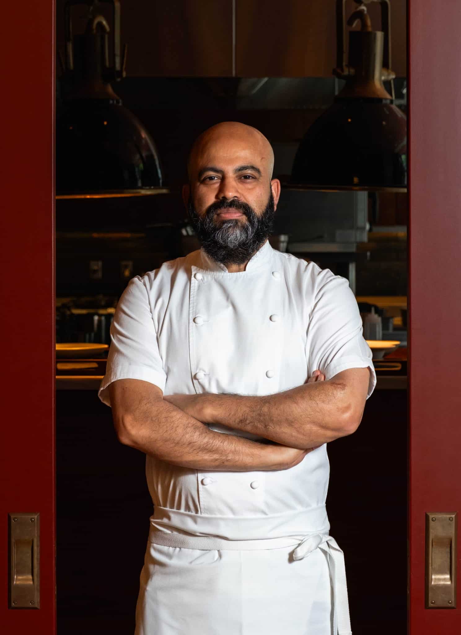 Chef Sujan Sarkar with a beard wearing a white chefs coat stands in a kitchen.