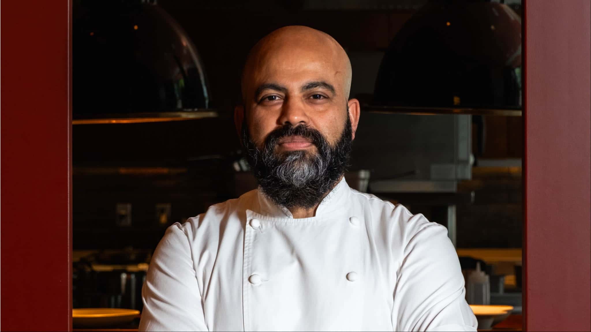 Chef Sujan Sarkar with a beard wearing a white chefs coat stands in a kitchen.