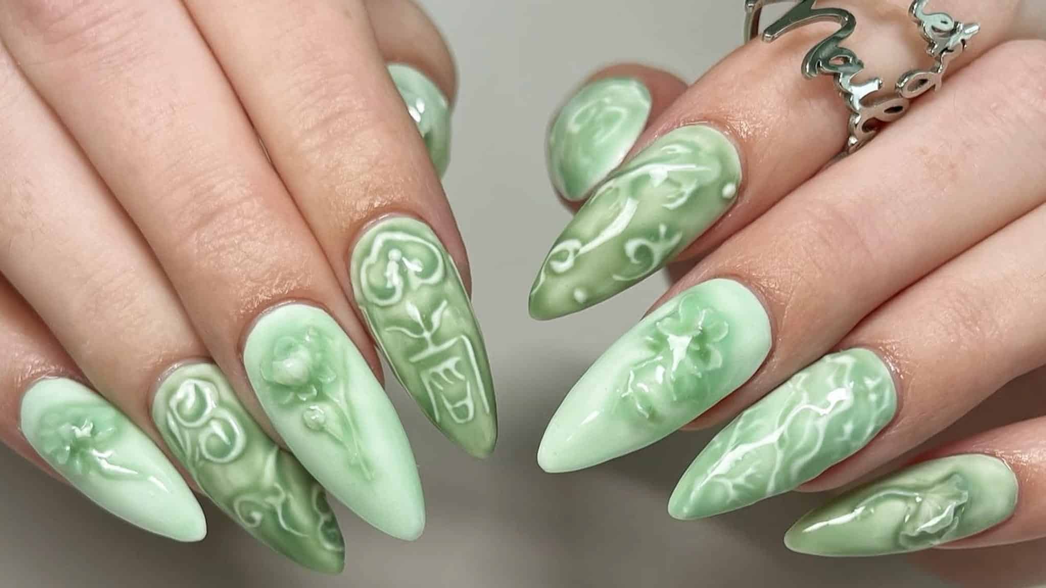 Hands showing stiletto-shaped nails with a sage green base and raised, white 3D floral and scrollwork designs.
