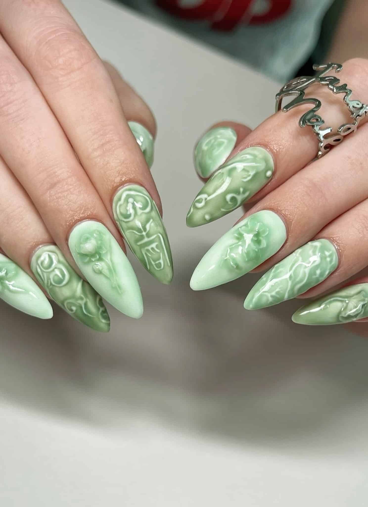 Hands showing stiletto-shaped nails with a sage green base and raised, white 3D floral and scrollwork designs.