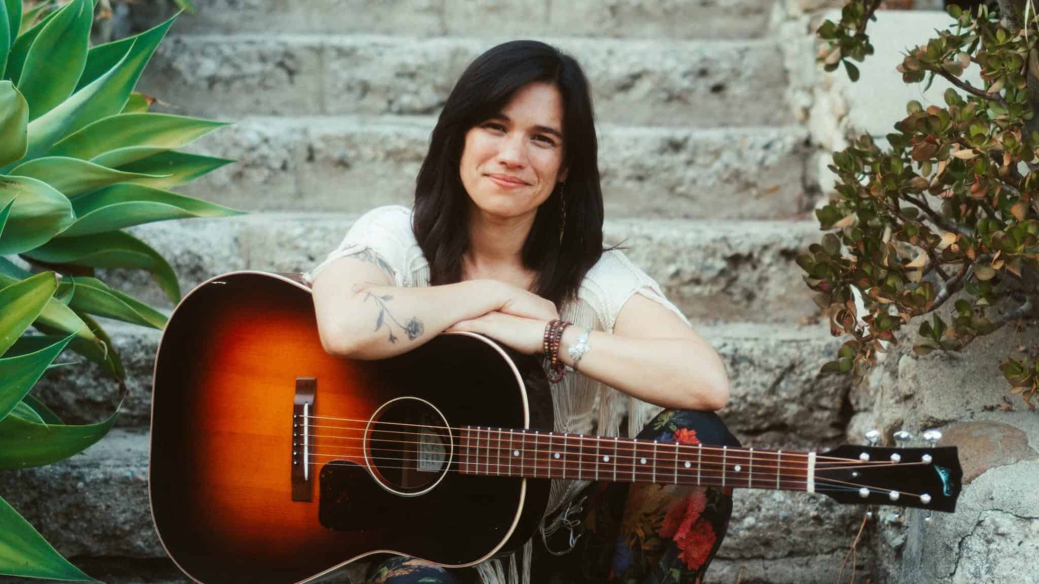 Paula Fong with long dark hair smiling while resting her arms on an acoustic guitar.