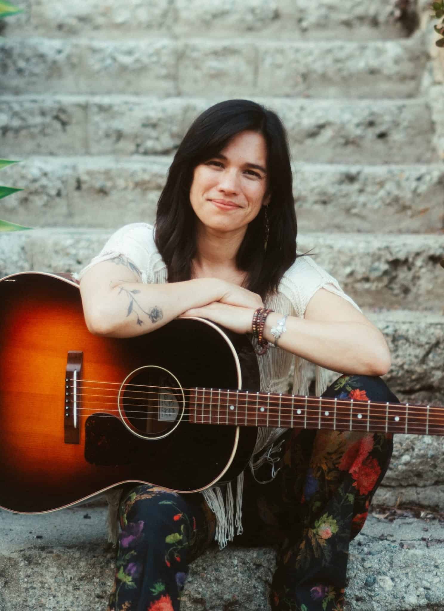 Paula Fong with long dark hair smiling while resting her arms on an acoustic guitar.