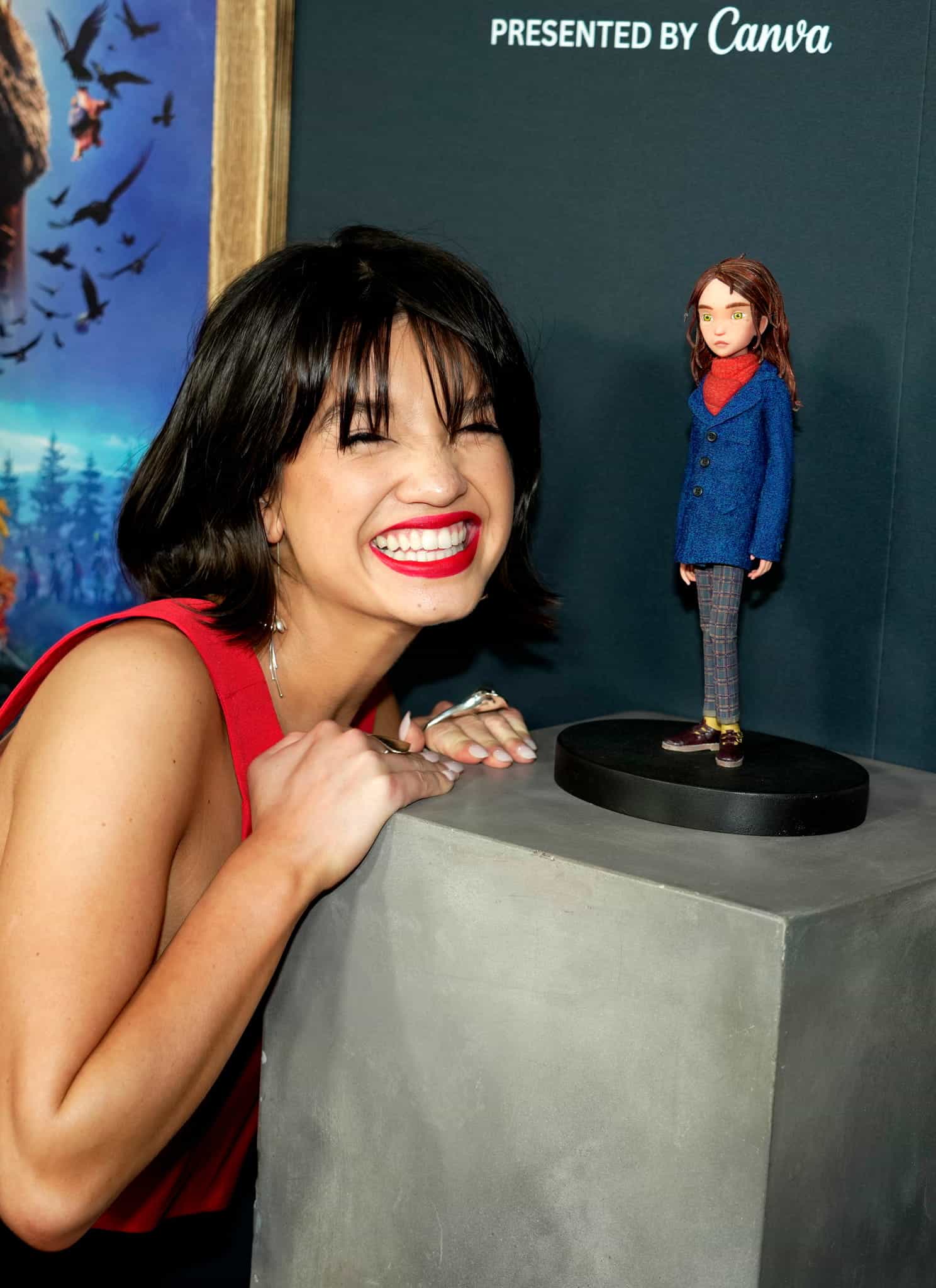 A smiling woman leans against a pedestal displaying a small figurine of the character Coraline.