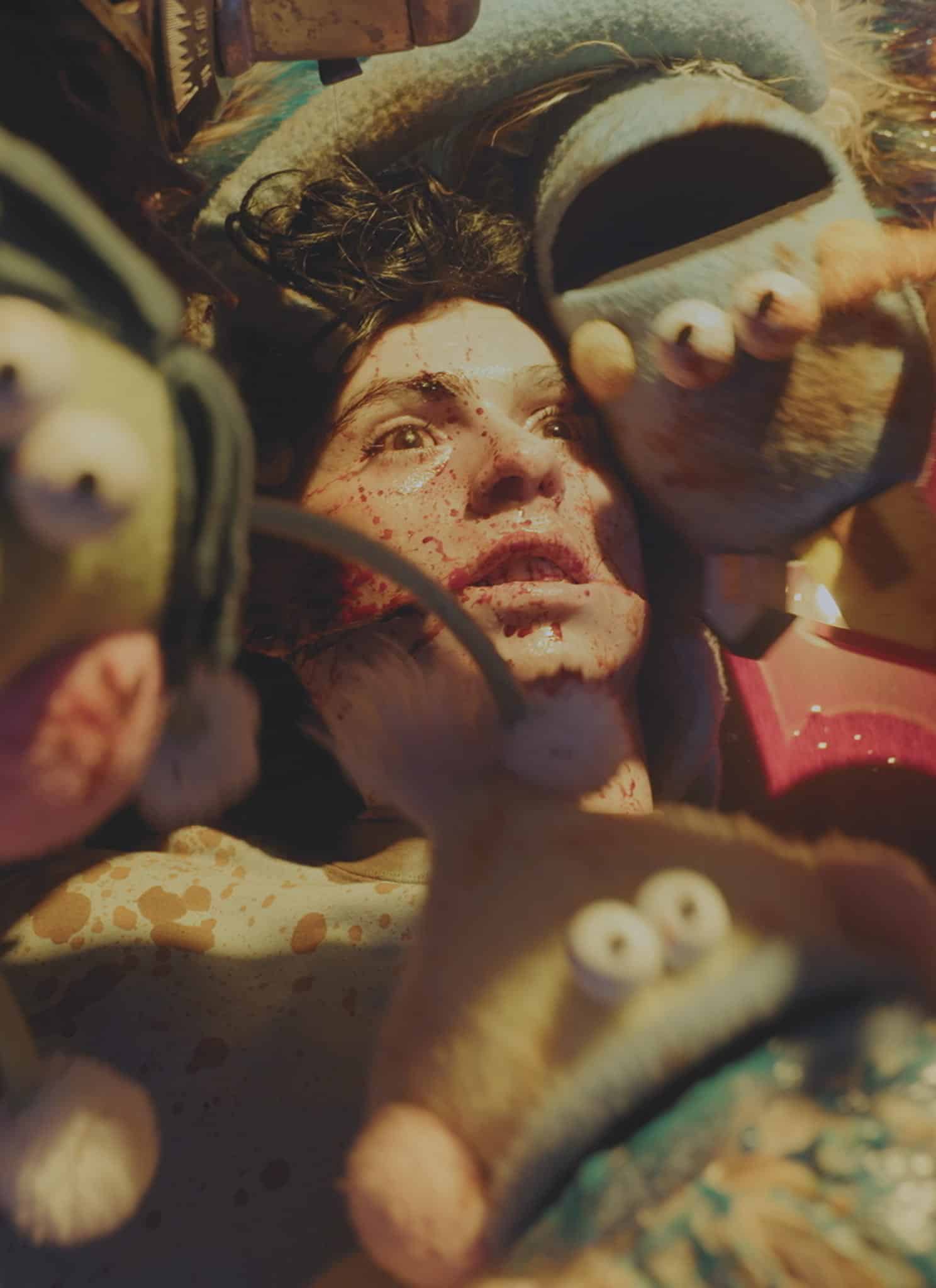 A person with blood-spattered skin lies on their back, surrounded by several stylized puppet creatures.