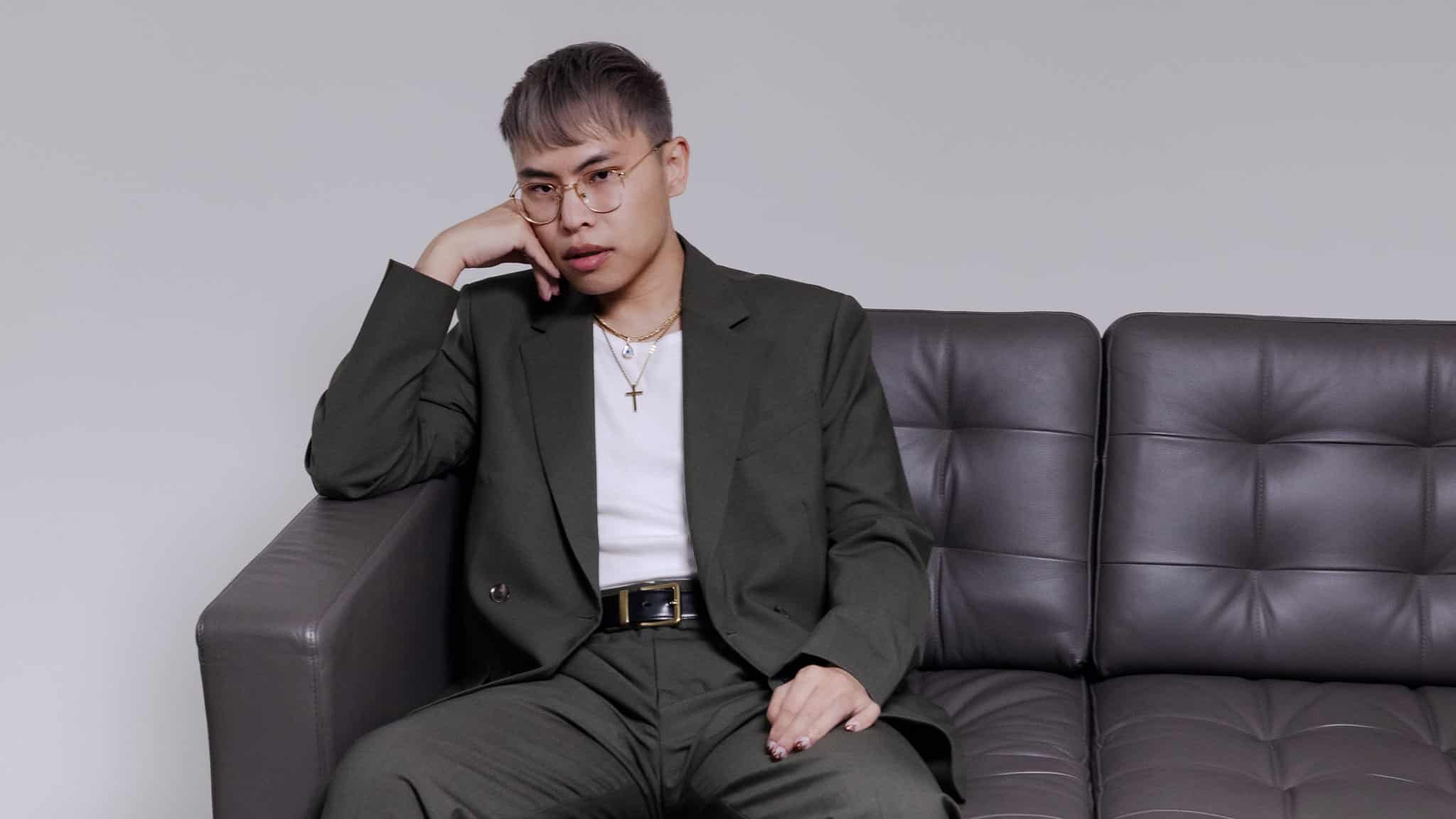 A person with short hair and glasses wearing a dark olive green suit and a white shirt sits on a grey sofa, resting their chin on their hand.