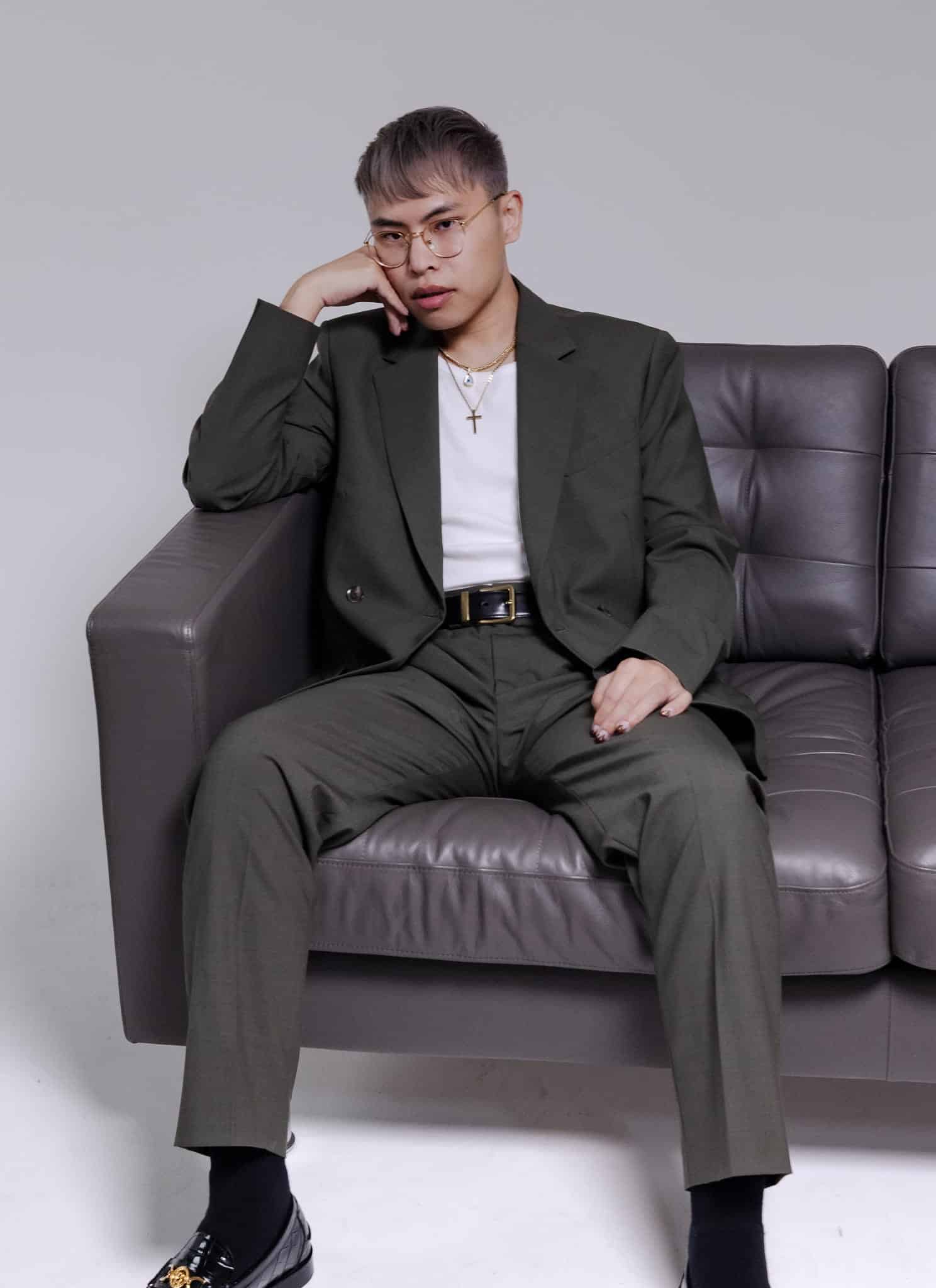 A person with short hair and glasses wearing a dark olive green suit and a white shirt sits on a grey sofa, resting their chin on their hand.