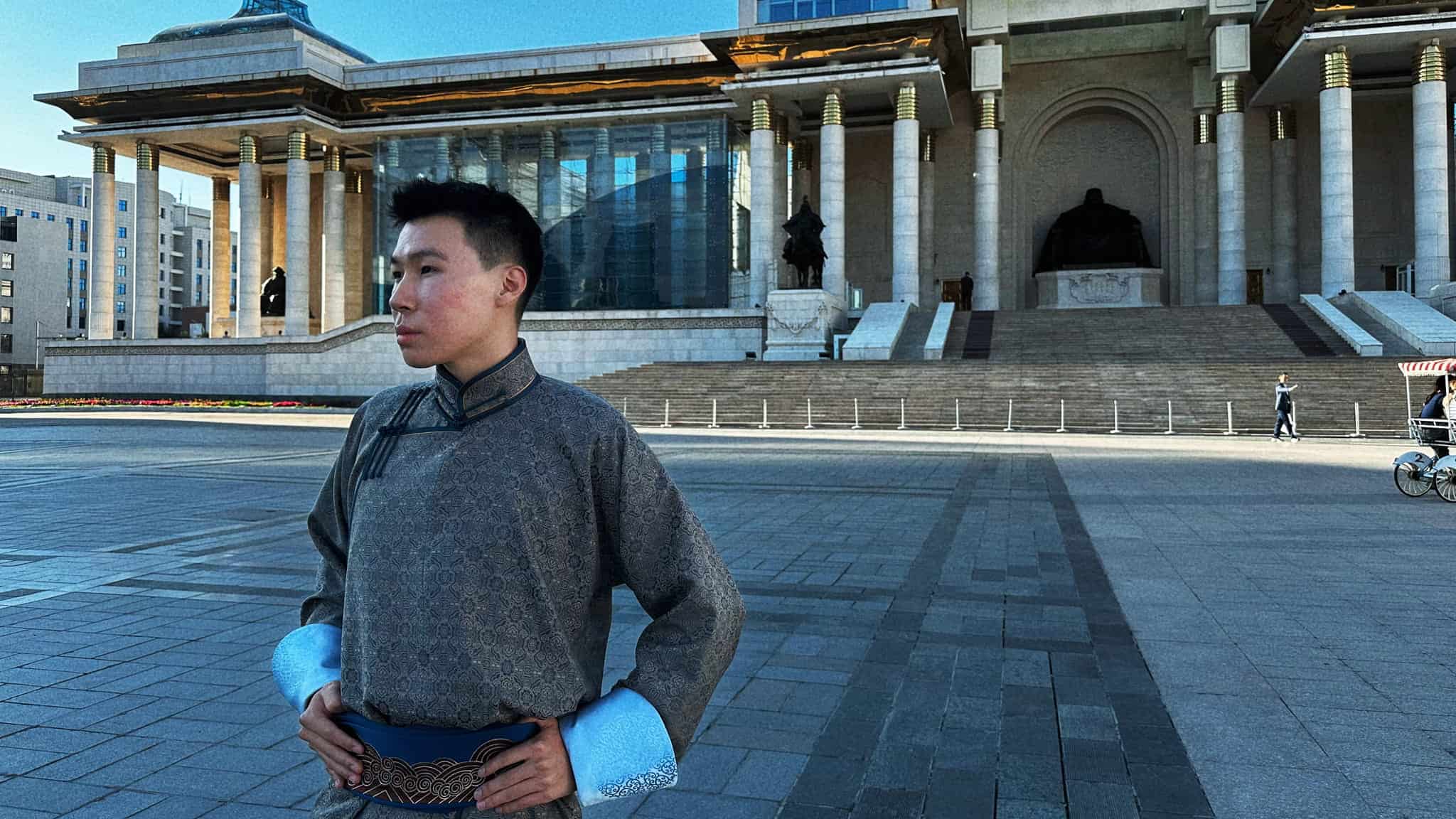 A man wearing a traditional Mongolian deel stands in a large paved square in front of the State Palace in Ulaanbaatar.