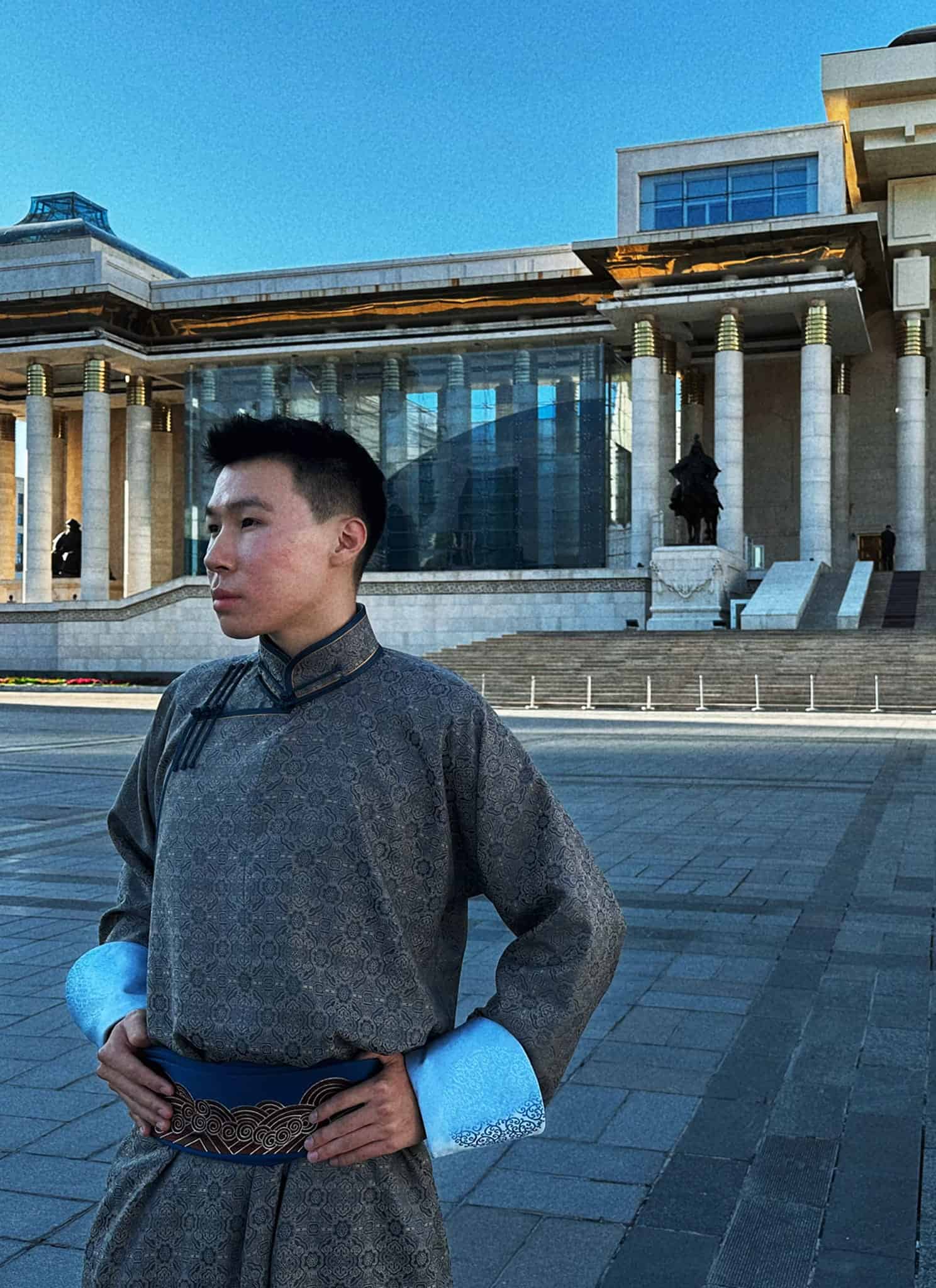 A man wearing a traditional Mongolian deel stands in a large paved square in front of the State Palace in Ulaanbaatar.
