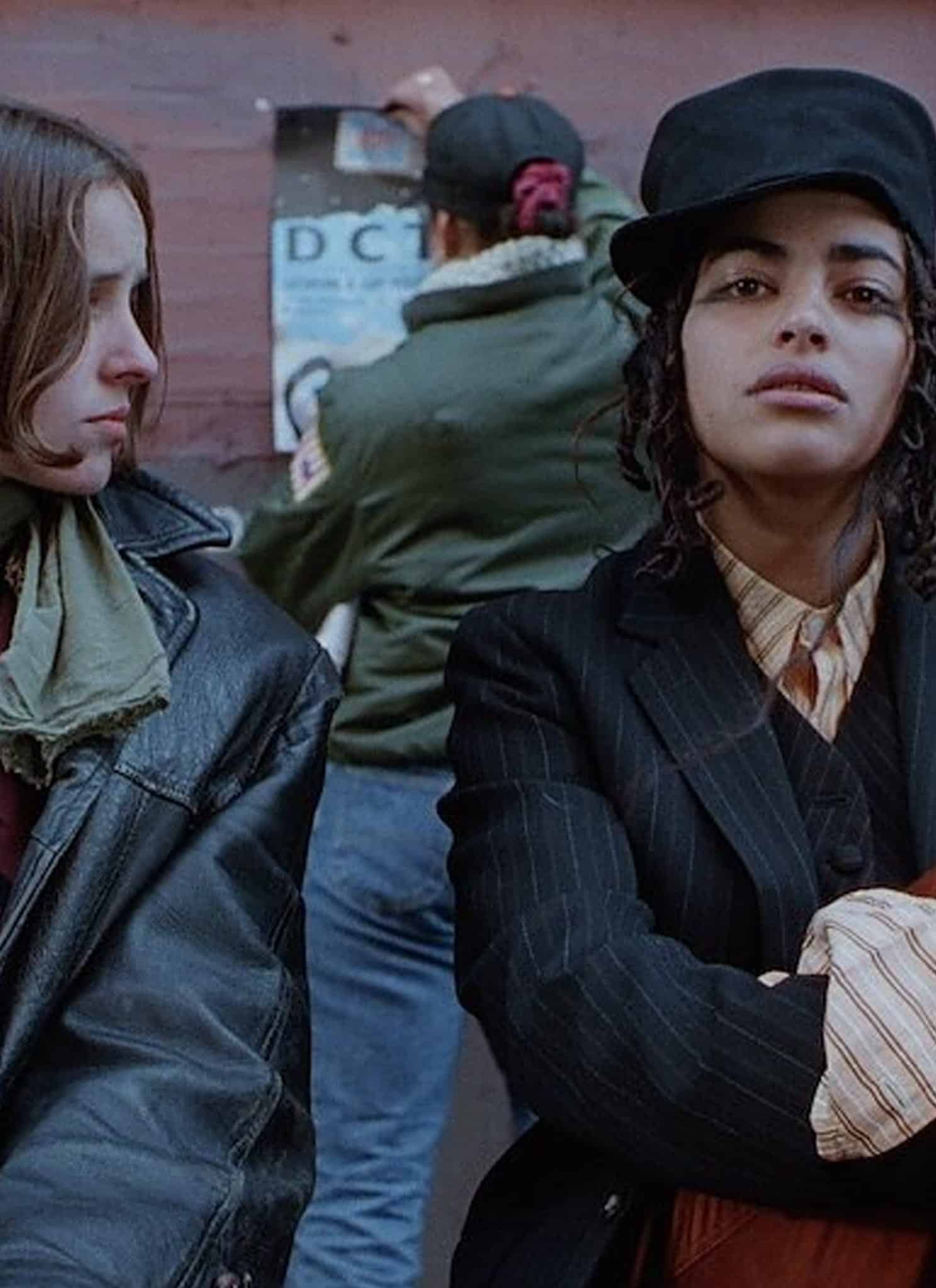 Two people sit in front of a brick wall while a third person pastes a poster behind them.