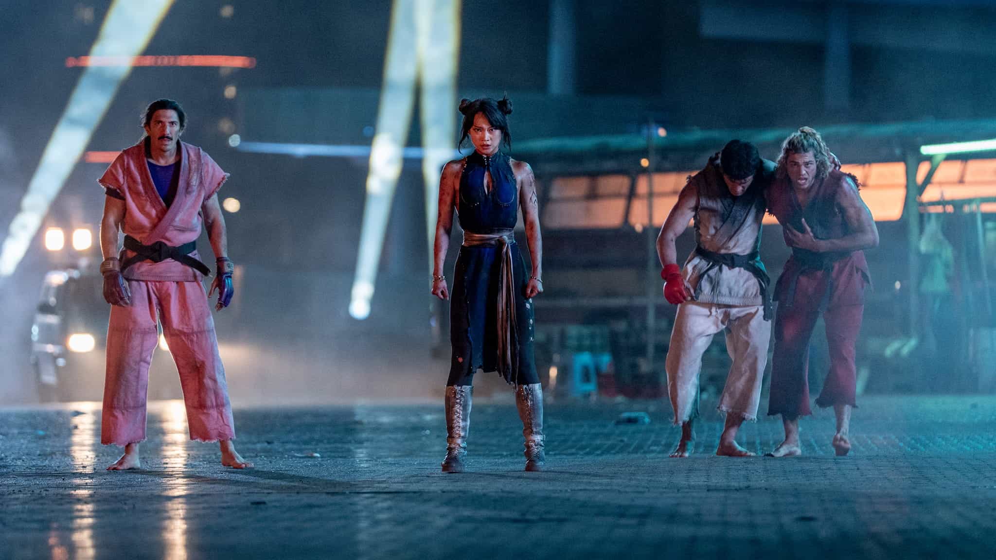 Four fighters in martial arts attire standing in a row at night, with one person in the center and two others supporting each other on the right.