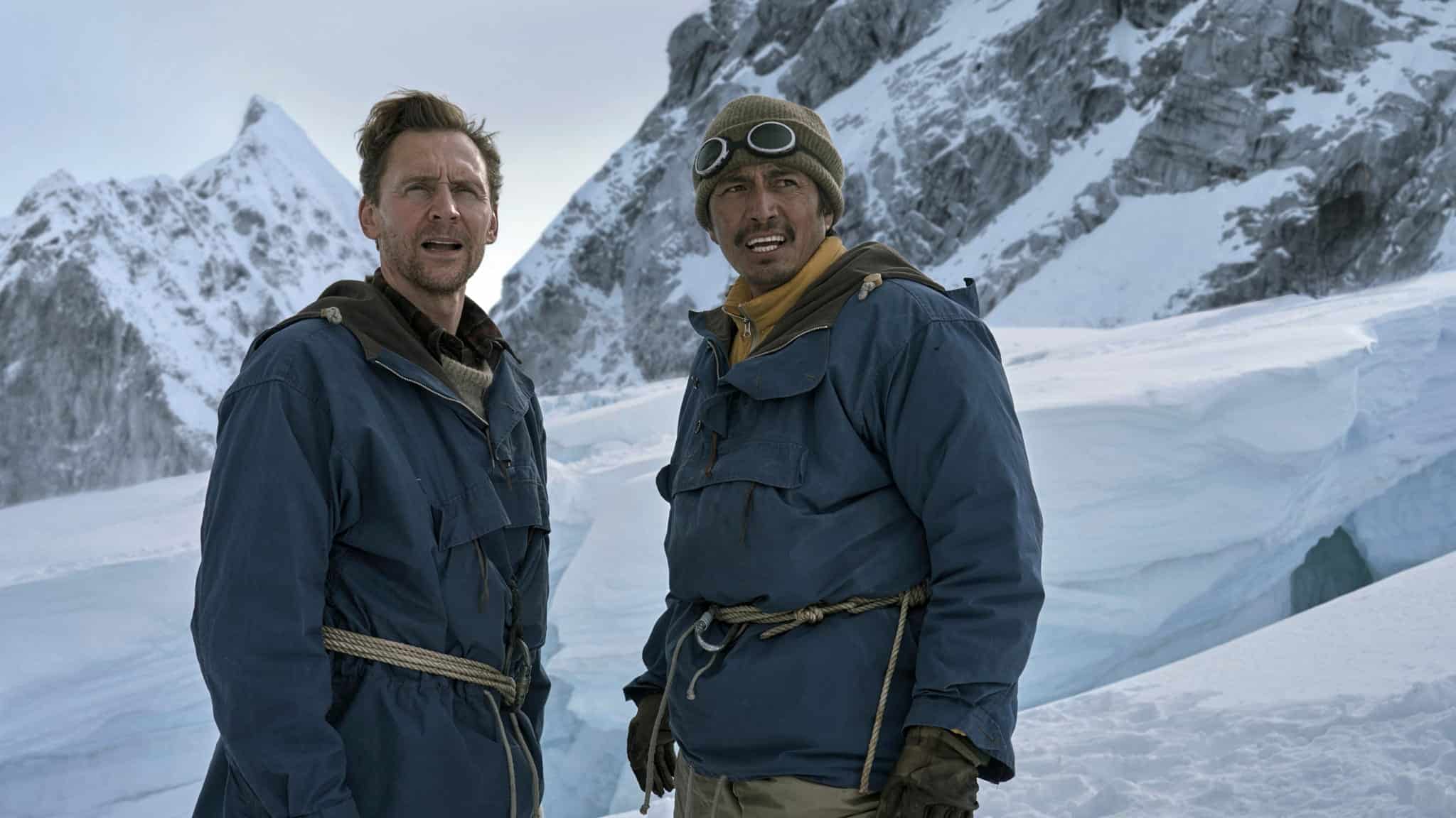 Two men in heavy blue parkas and climbing gear stand on a snow-covered mountain.