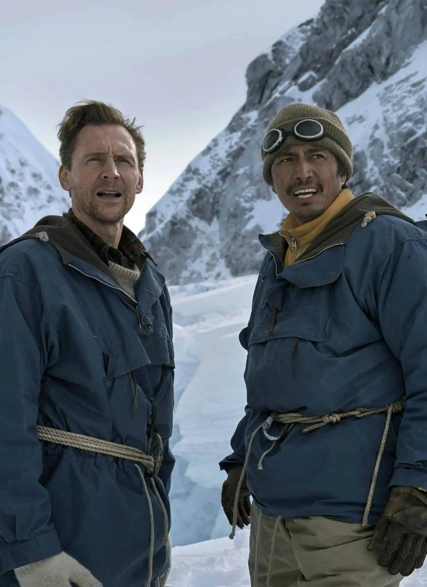 Two men in heavy blue parkas and climbing gear stand on a snow-covered mountain.