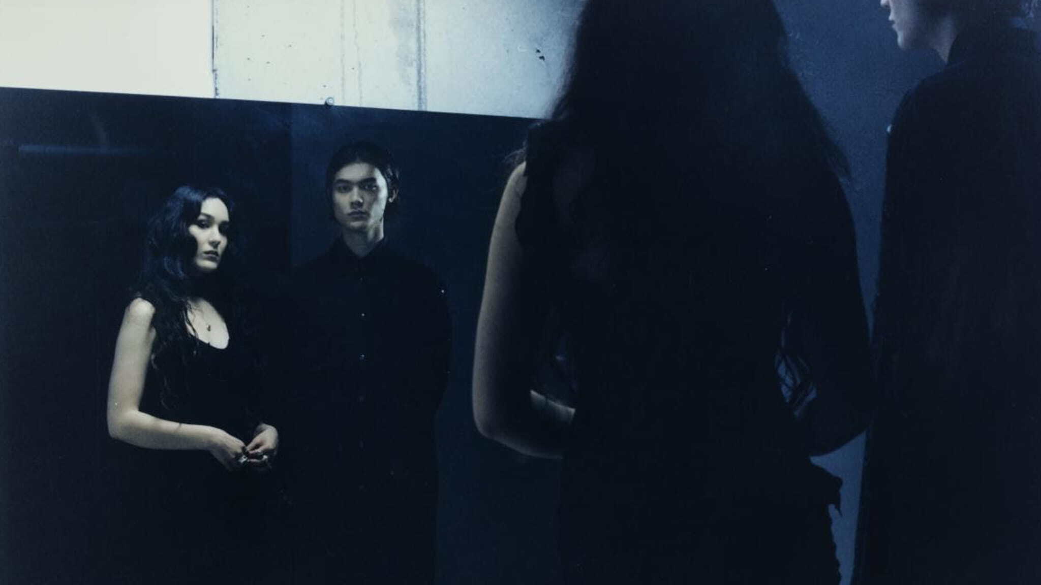 A woman and a man in dark clothing stand before a large mirror, with their reflections visible.