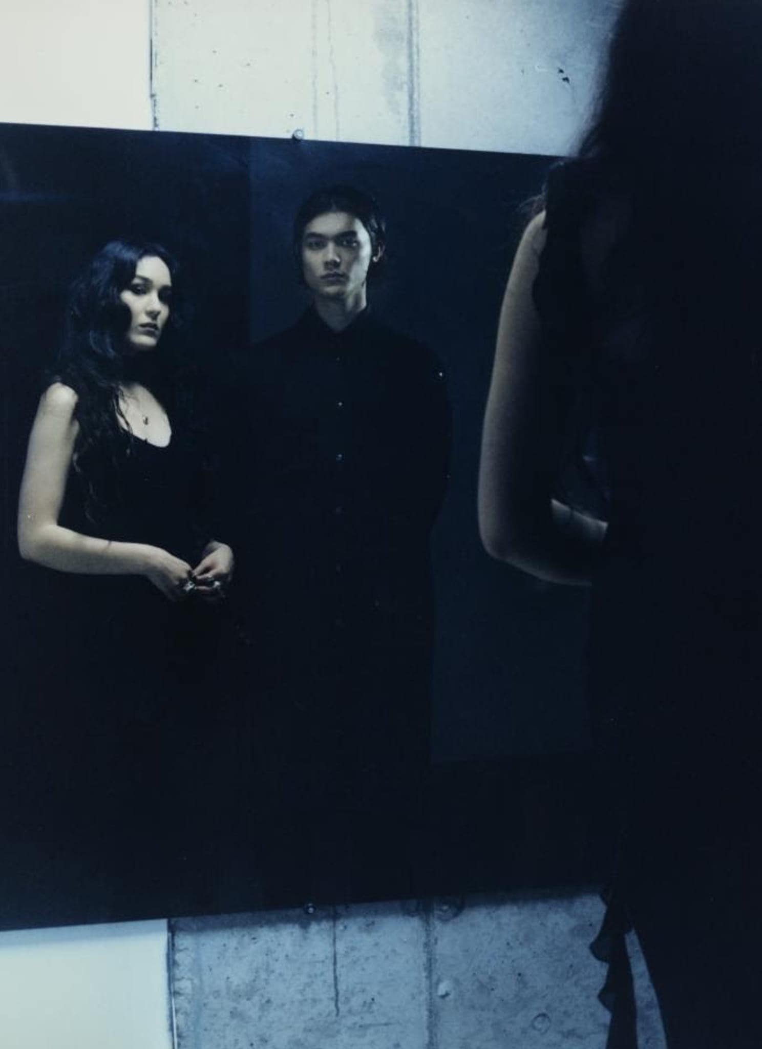 A woman and a man in dark clothing stand before a large mirror, with their reflections visible.