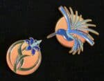 Two round enamel pins on a black background; one features a blue and green hummingbird whose wings are book pages in front of a gradient orange circle, the other shows a purple flower with green stems and a fountain pen stamen against a similar gradient orange circle.