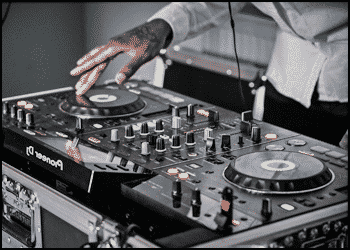 A person wearing a light-colored shirt operates DJ Controllers, adjusting knobs and spinning a jog wheel during a music mix.