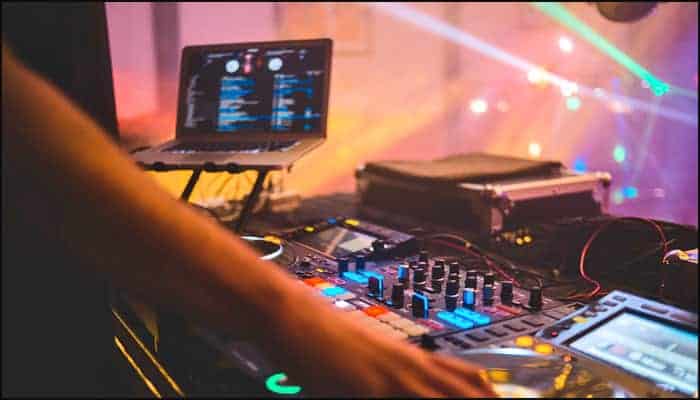 A DJ operates a mixing console and laptop set securely on DJ Laptop Stands, with colorful lights and a blurred background, suggesting an active nightclub or party scene.